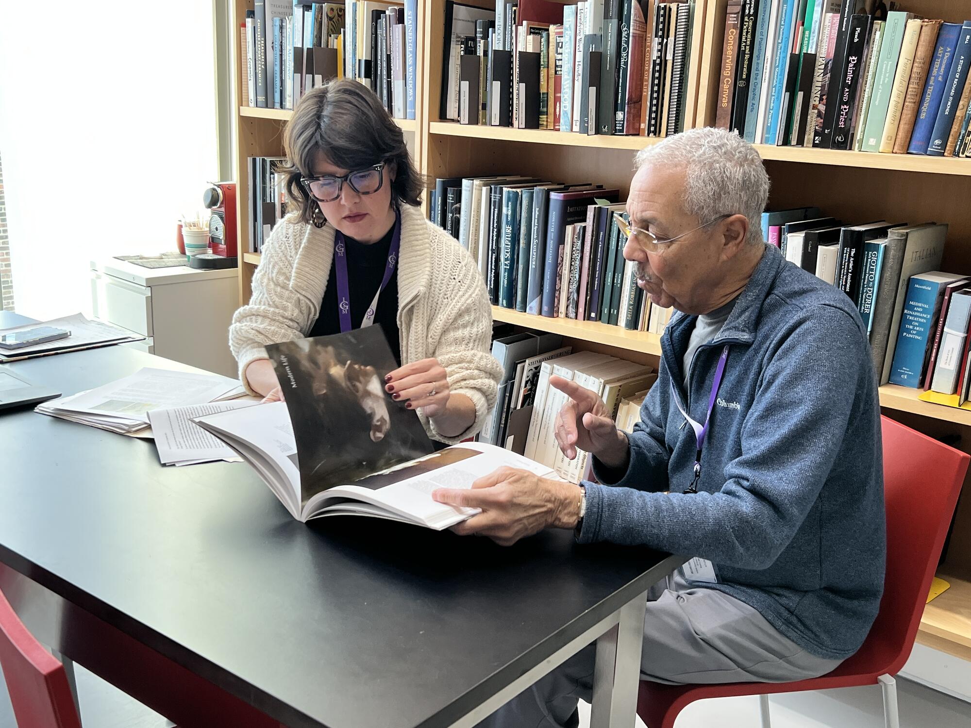 A caucasian woman with glasses looking through a book with an African American man with grey hair and glasses with a book-filled case behind them. 