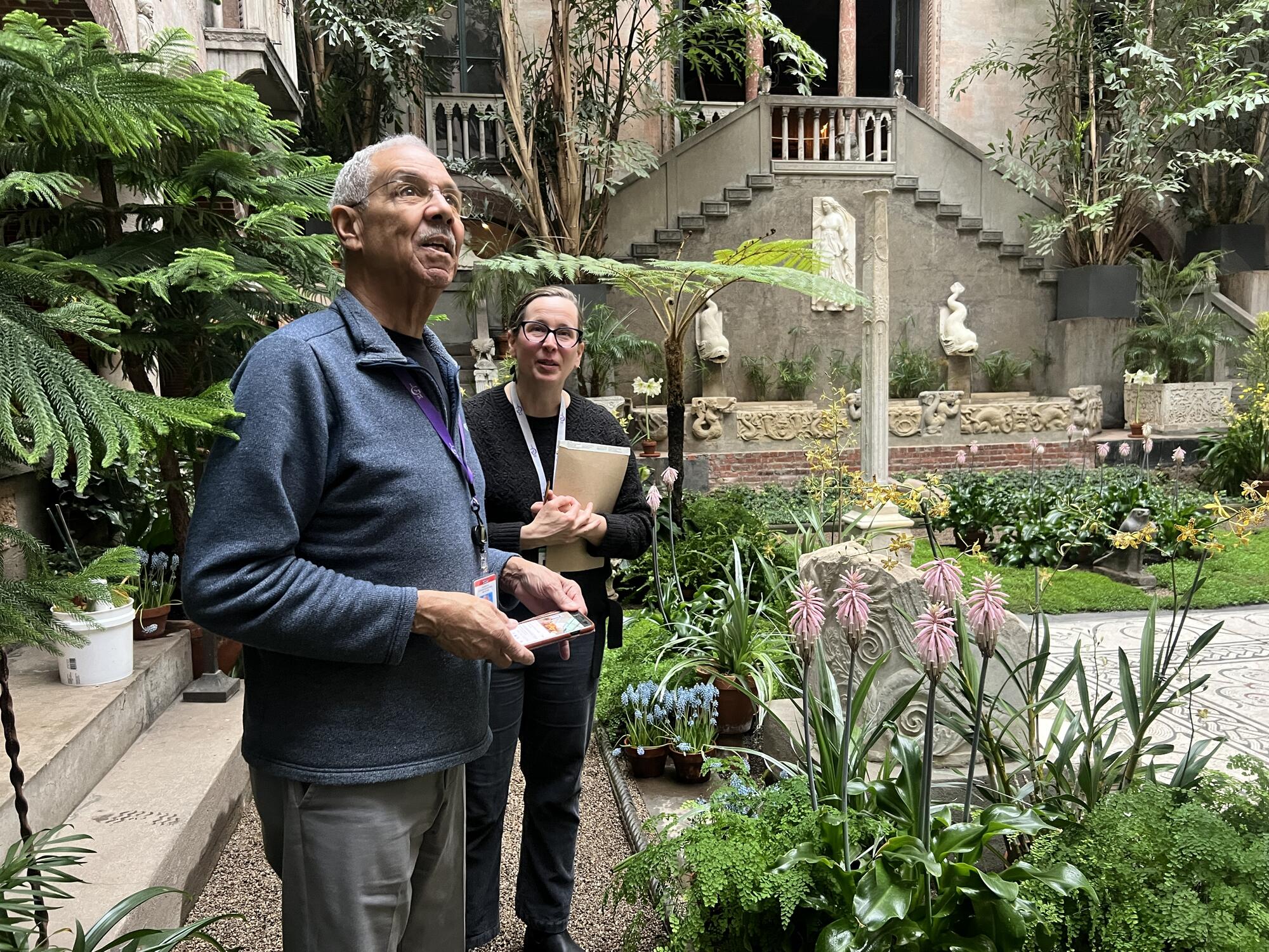 African American man with grey hair standing in the courtyard with a causacian worman holding a notebook.  