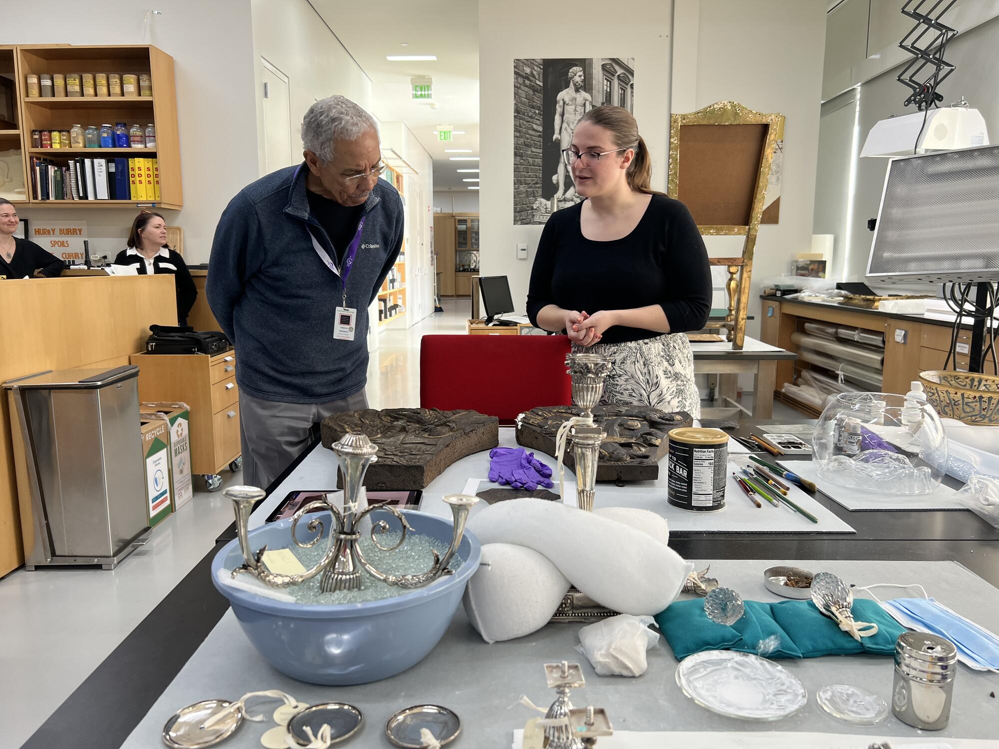 African American man with grey hair bends down to look at items on a table top with a Caucasion woman standing next to him. 