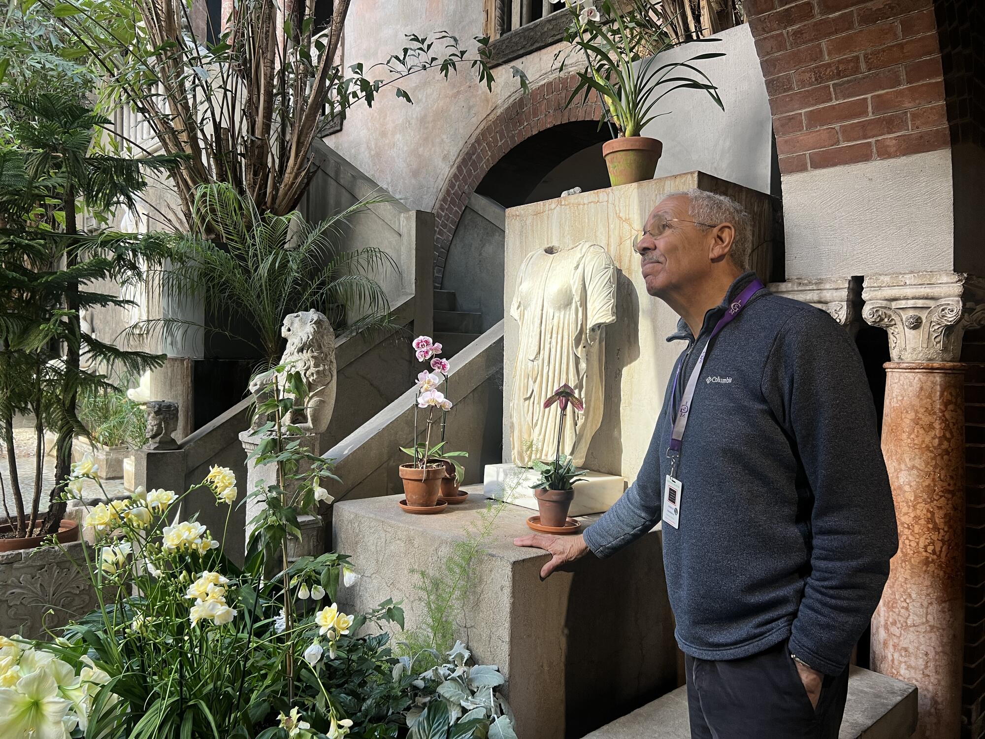 An African American man with grey hair looking up towards the ceiling in the courtyard surround by lush, green plants. 