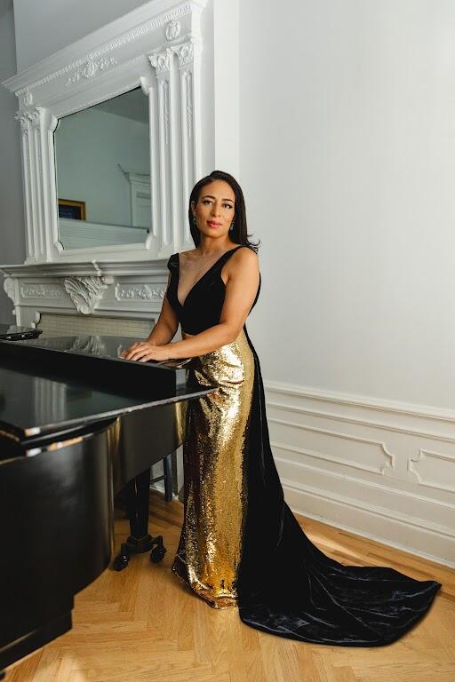 Woman with long dark hair wearing a gold and black sleeveles gown standing next to a grand piano with a white wall, fire place and mirror behind her.  