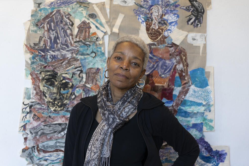 African American woman with short cropped grey hair with a black top and scarf tied around her neck standing in front of a colorful painting with mermaids and the ocean. 