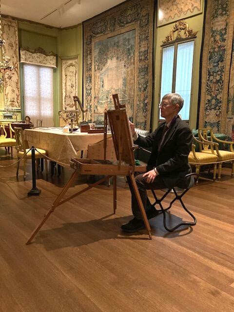 Woman with short grey hair sitting on a chair drawing on an easel in an ornately decorated room.  