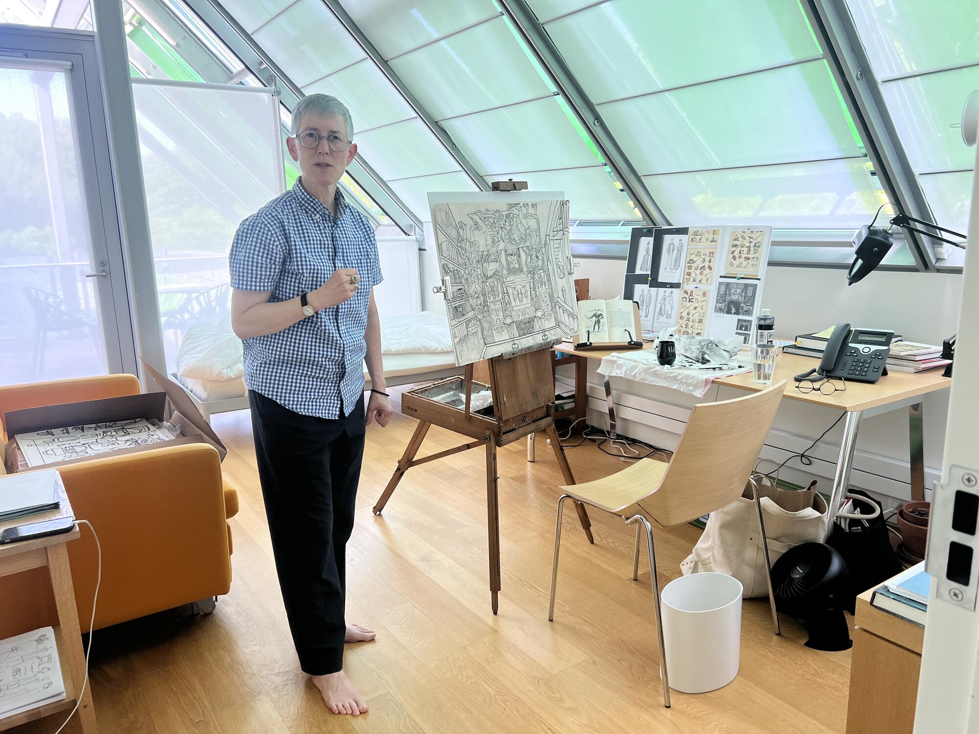 Woman with short grey hair wearing an oversized blue shirt and black slacks standing in a sunny studio space