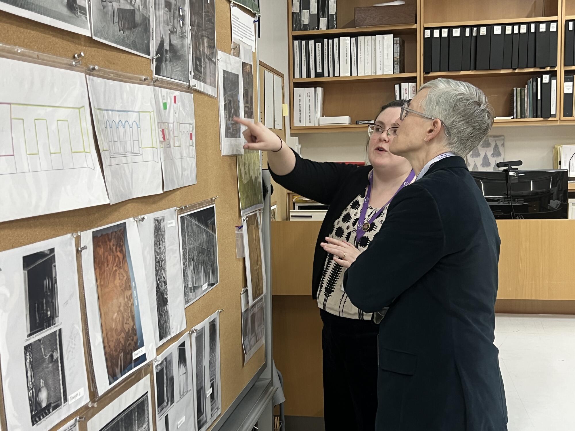 A woman with short grey hair looking at textile samples on a bulletin board with another woman pointing to a sample