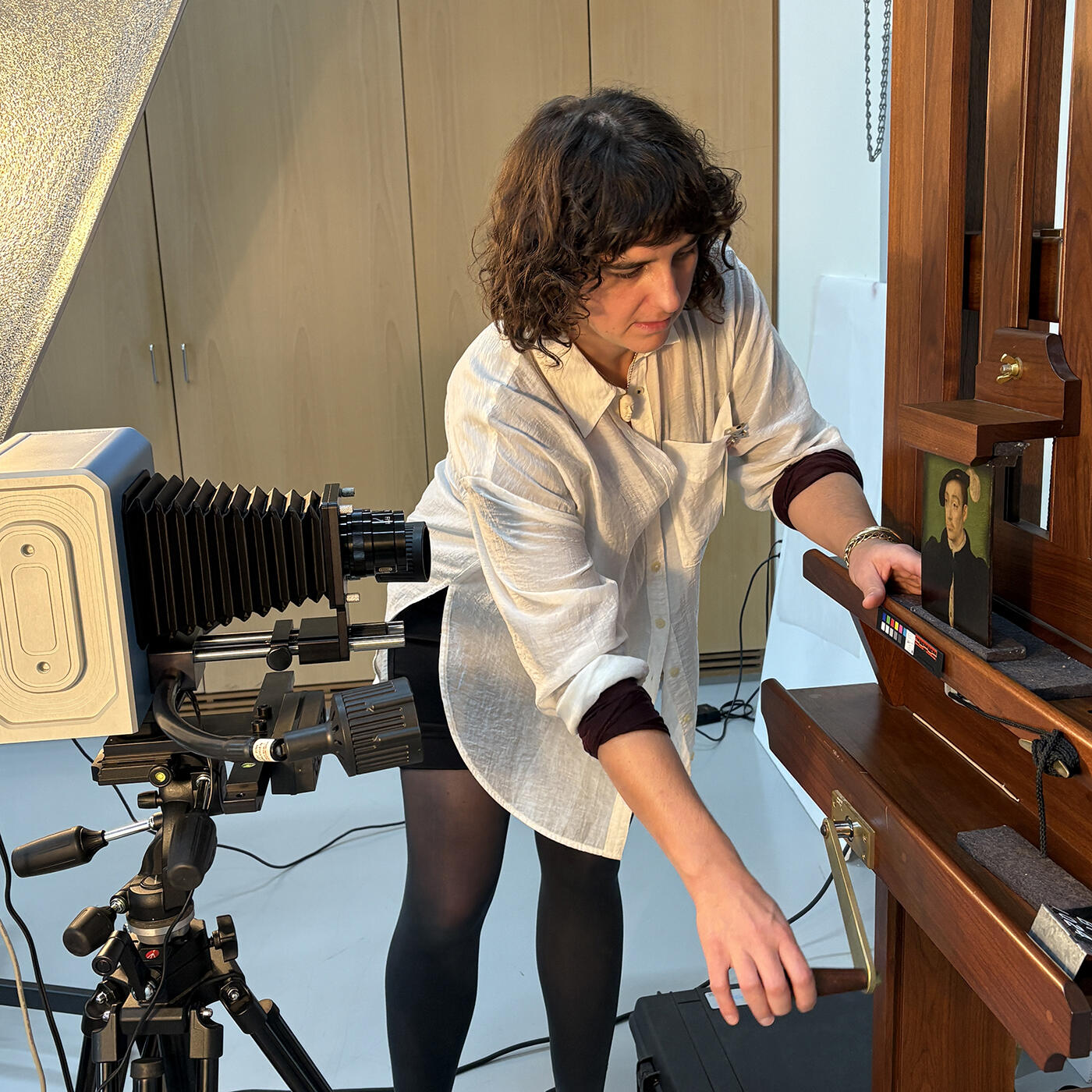 A conservator adjusts the height of an easel supporting a small portrait of a man in front of an IRR camera on a tripod.