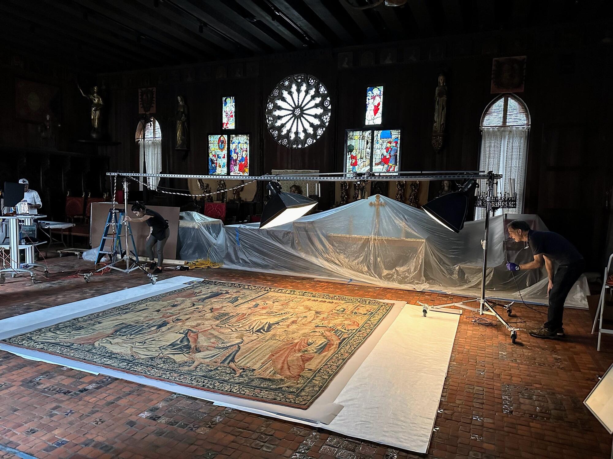 A tapestry laying on the floor with lights and a camera suspended above it from two steel rails; a person on either side moving the rails to a new position over the tapestry.