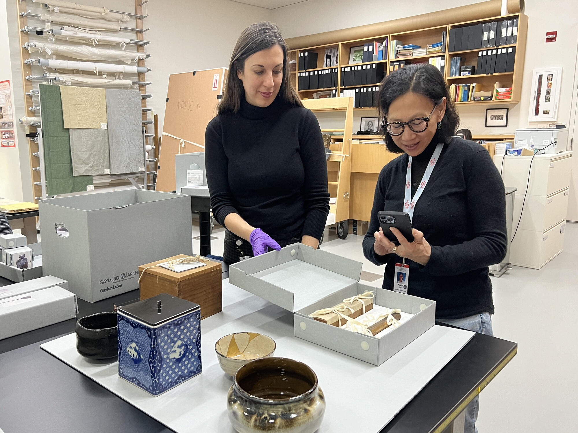 Two women looking at tea set items from an archival box laid out on a desktop. 