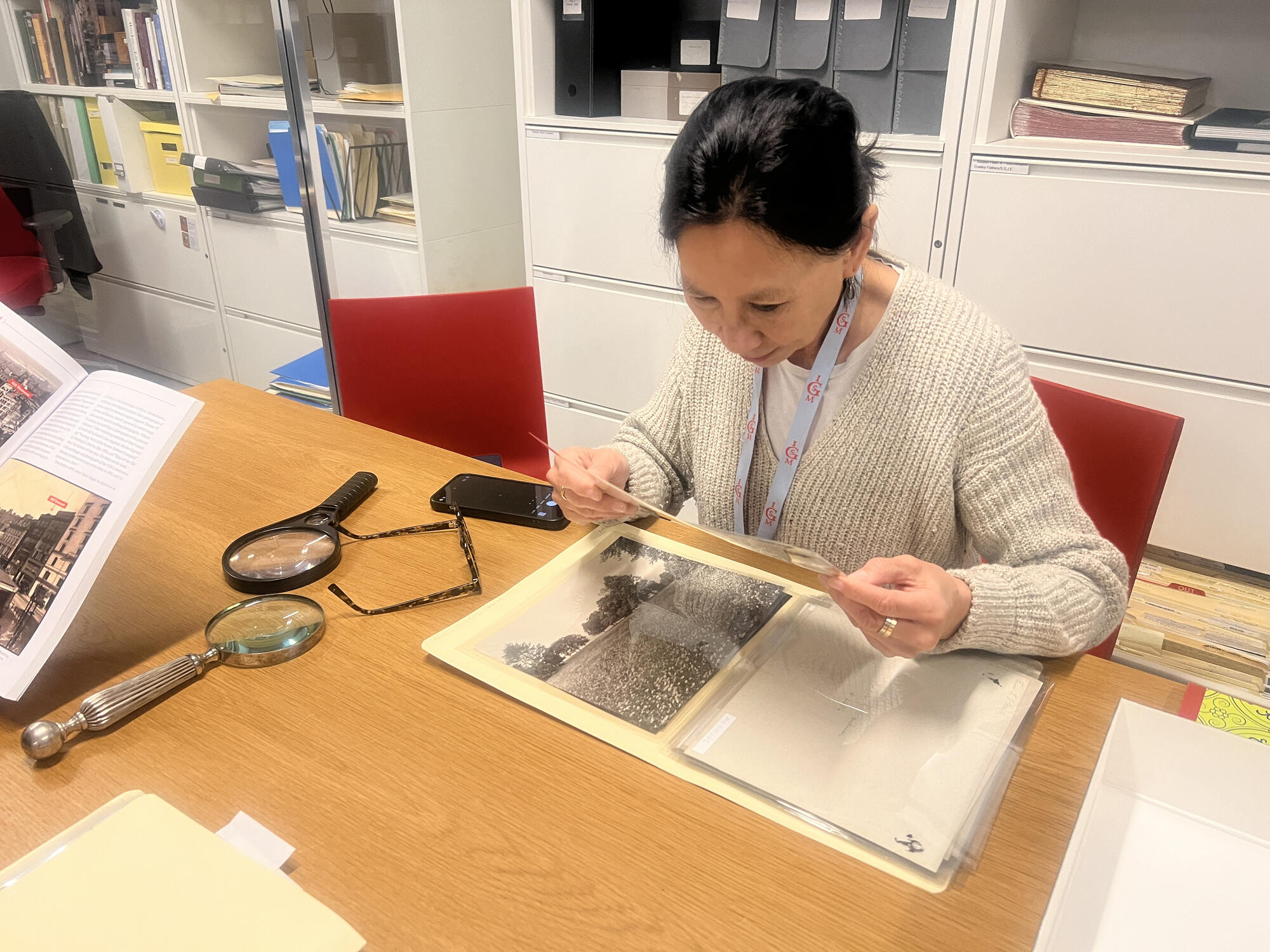 A woman sitting at a desk intently studies a page from a scrapbook.  