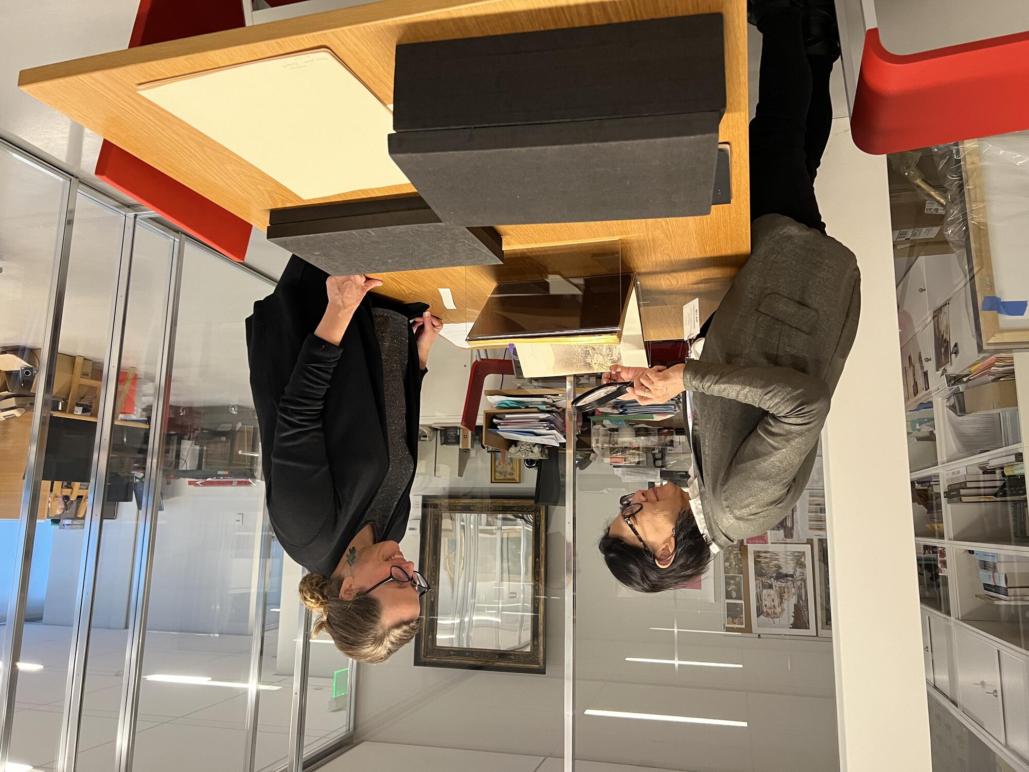 Two women standing over a desk looking at a book from an archive box and one woman is looking at its pages with a magnifying glass. 