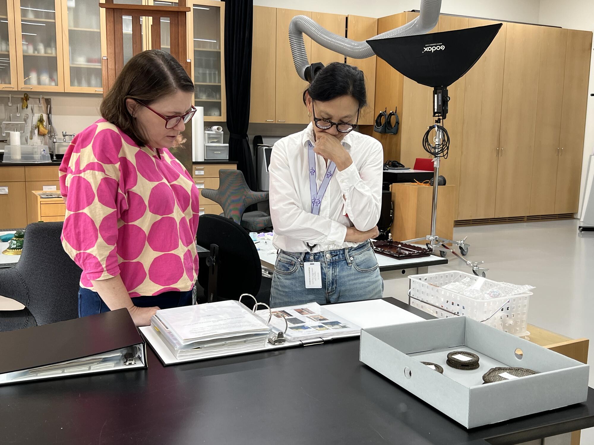 Two women looking intently at a notebook with slides on top of a desk in a room that looks like a laboratory. 