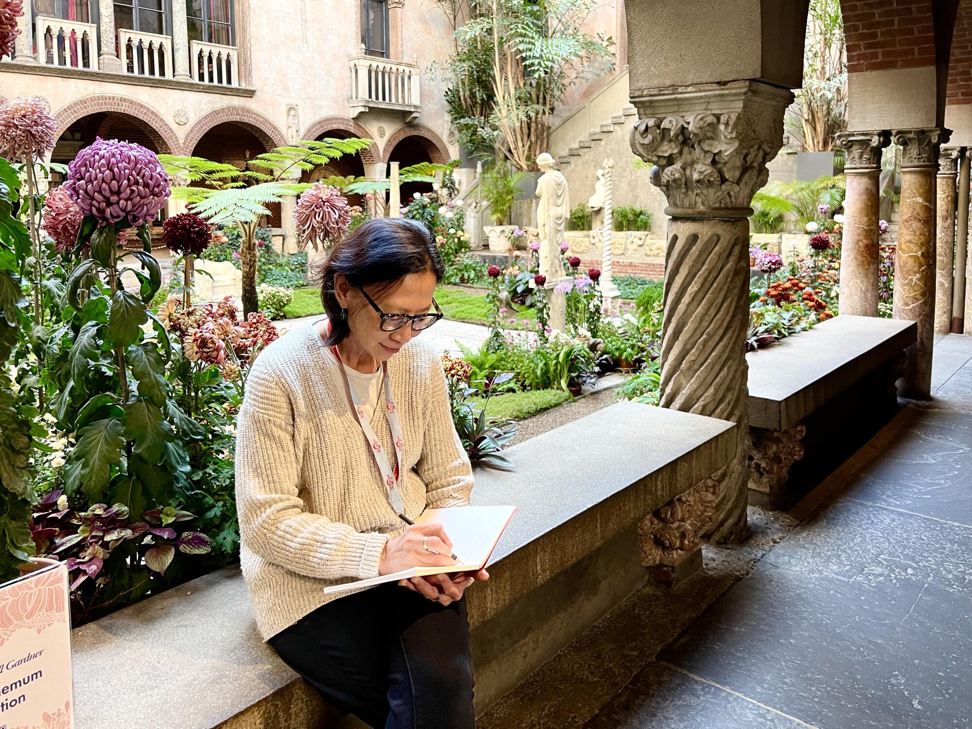 A woman sitting on a bench in the courtyard with a notebook/sketchbook on her lap. 