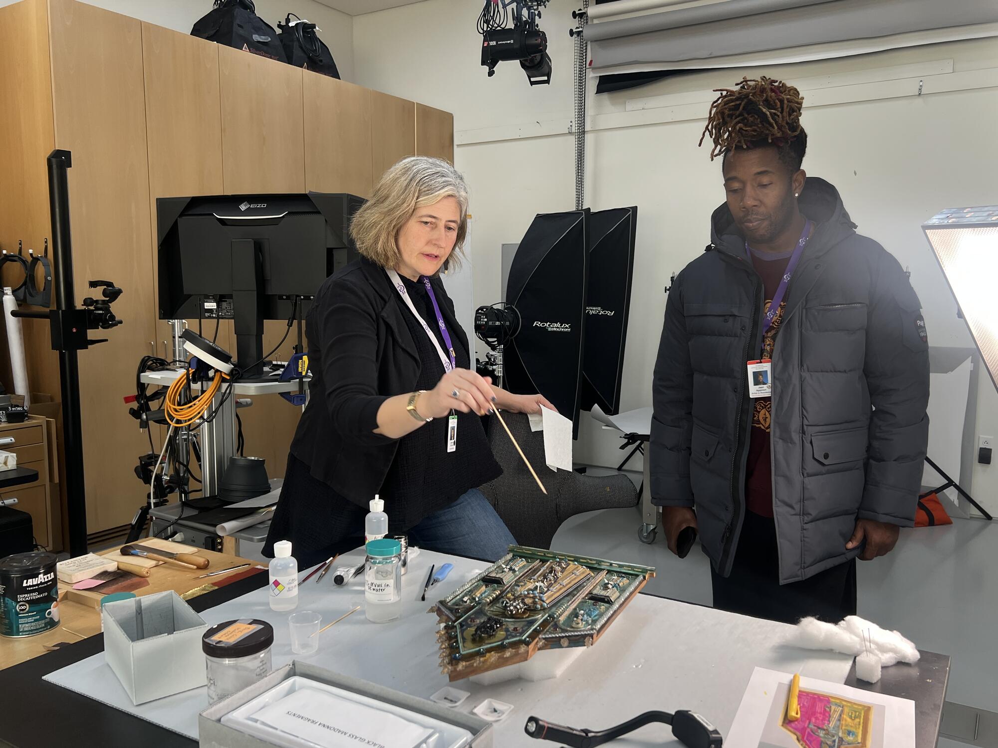 An African American man stands with a white woman as she points to an ornate object on a table with cleaning materials near by. 