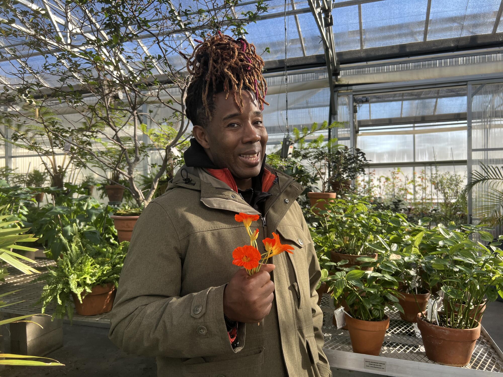 An African American man with braids piled on his head, holding nasturtium flowers while standing in a greenhouse. 