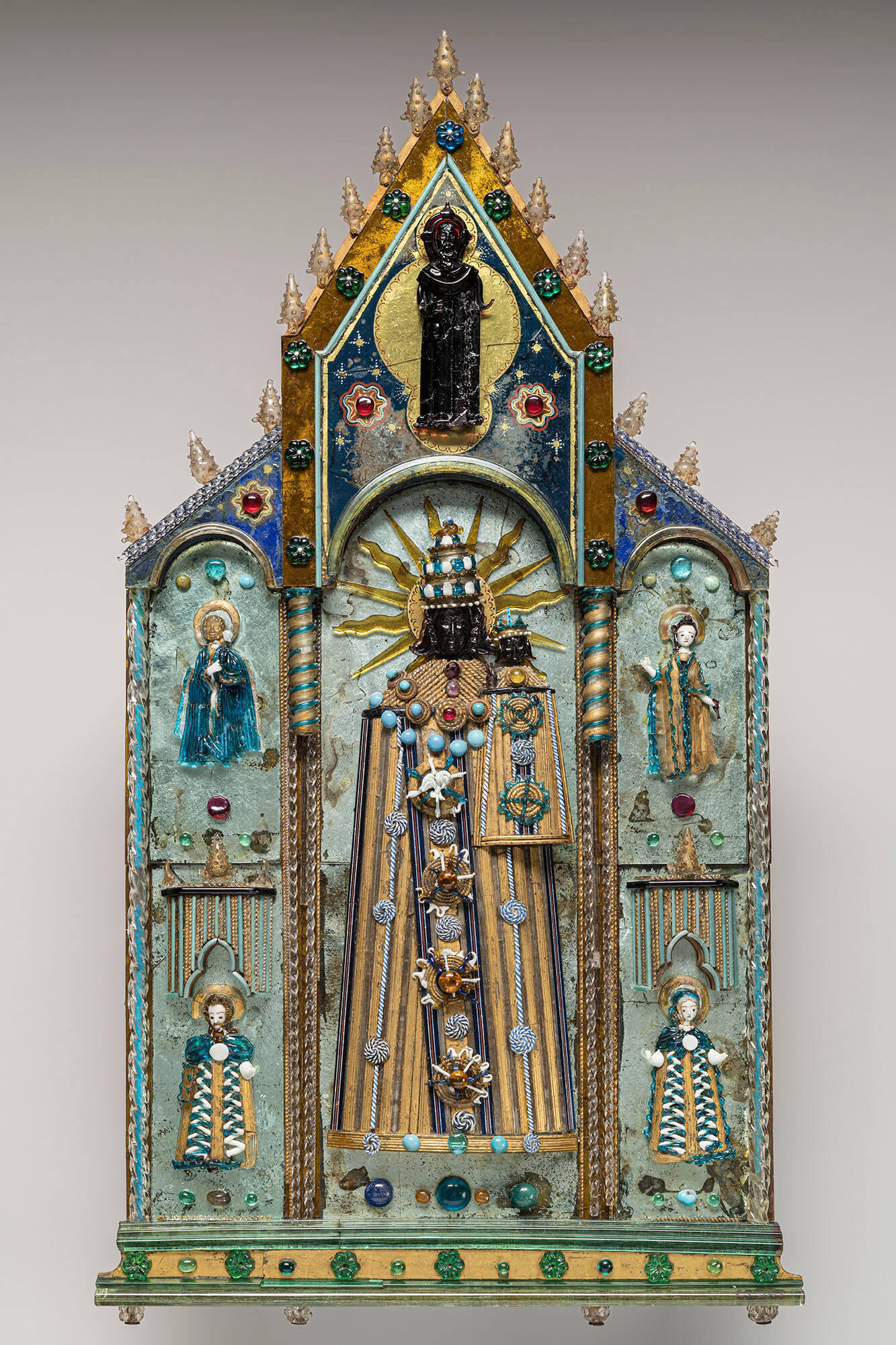 A panel of glass work. A Virgin and Child with black faces is in the center. They are surrounded by four saints with white faces. On the pinnacle is a depiction of God the Father in black glass.