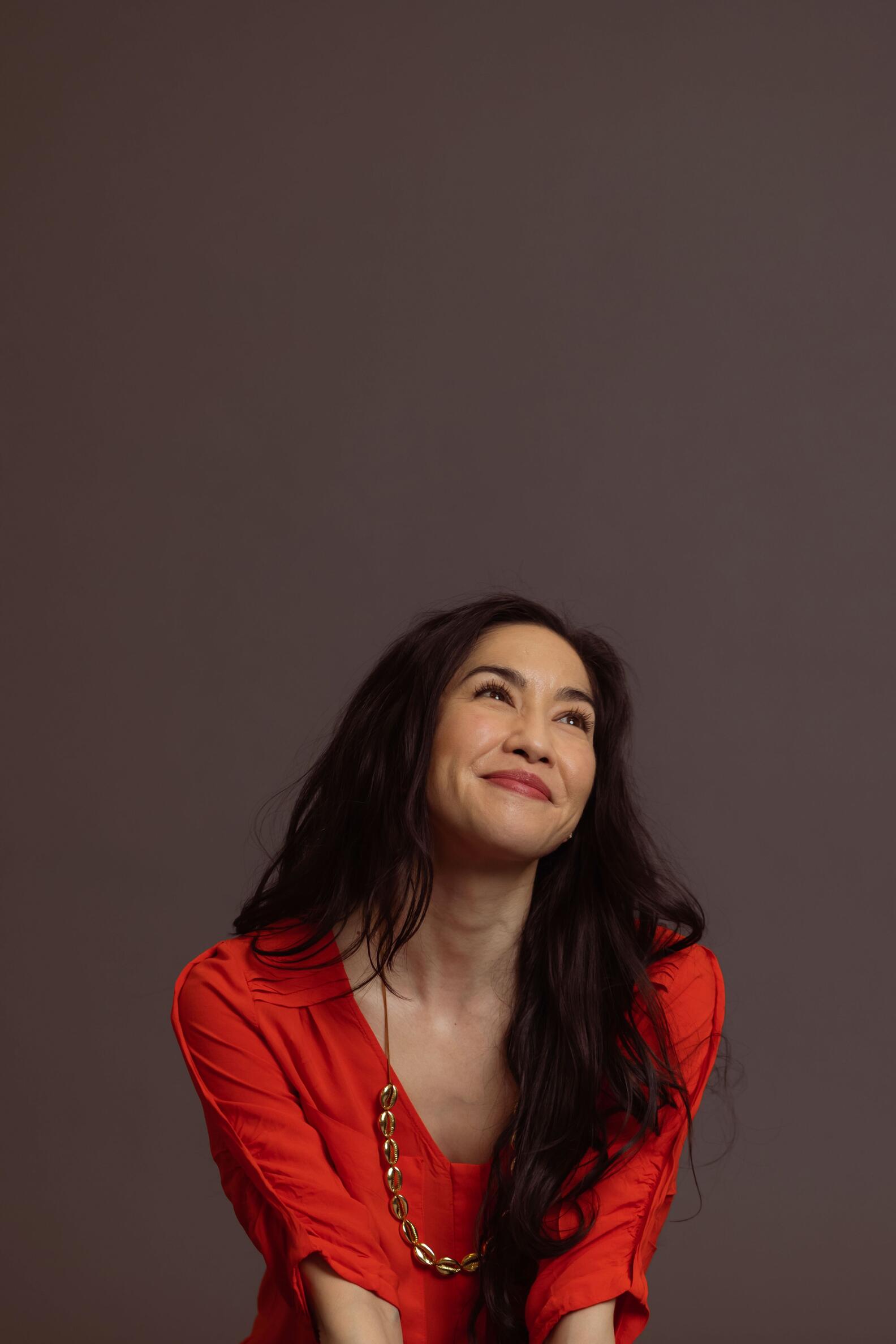 Photo of an Asian American woman with long black hair wearing an orange top loooking upwards, smiling