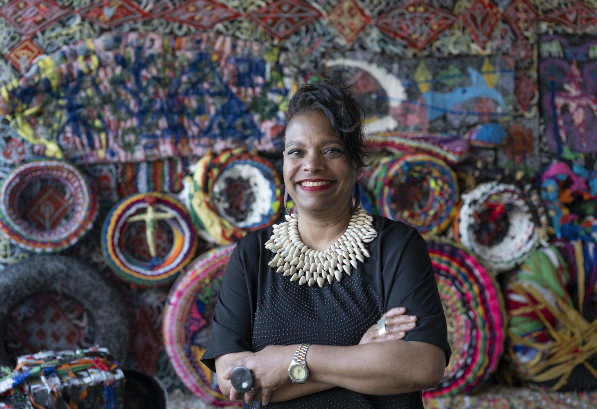 Fair skin African American woman with dark hair pulled up with a black top with a large, gold, necklace standing in front of a colorful wall with many circles of artwork. 