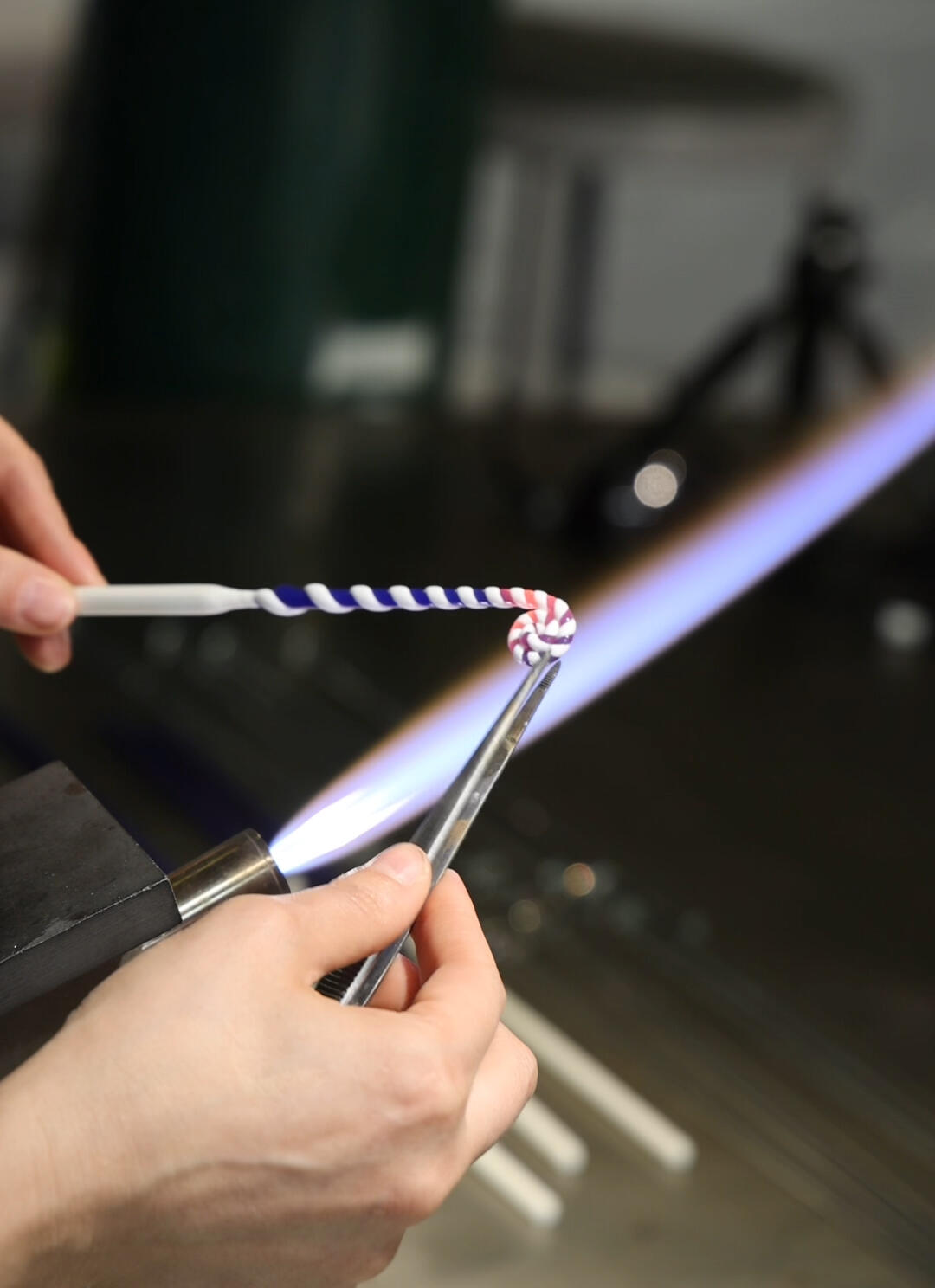 Two hands use a tool to manipulate blue and white glass under a flame.