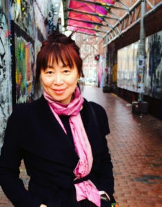 Asian woman with long dark hair with reddish highlights pulled up into a messy bun wearing a black jacket and pink scarf standing in an alley way with art adorning the walls. 