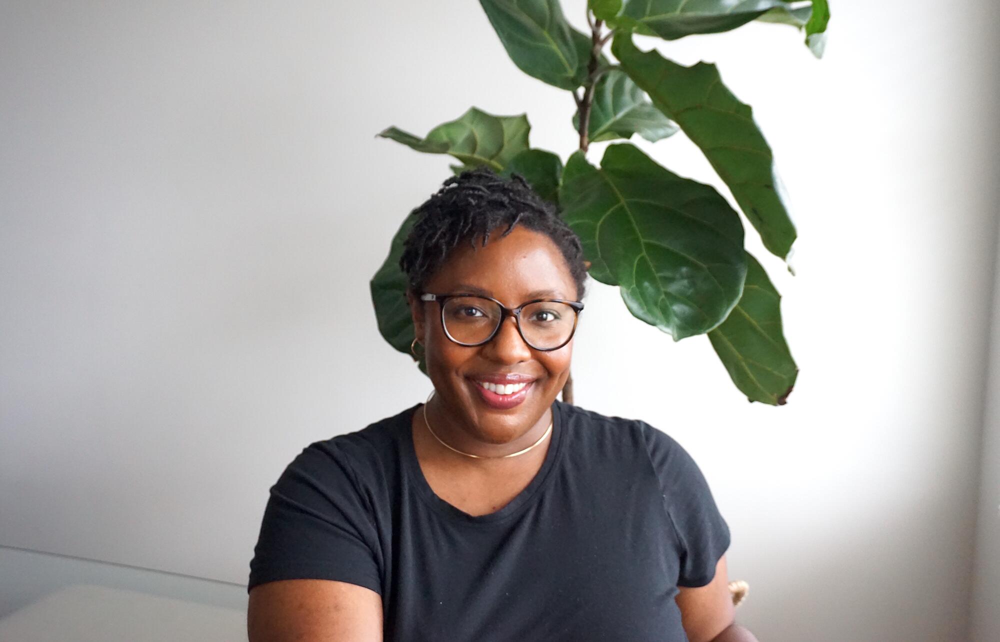 Young African American woman with short dreadlocks wearing a black t-shirt and glasses sitting in front of a large plant and white wall. 