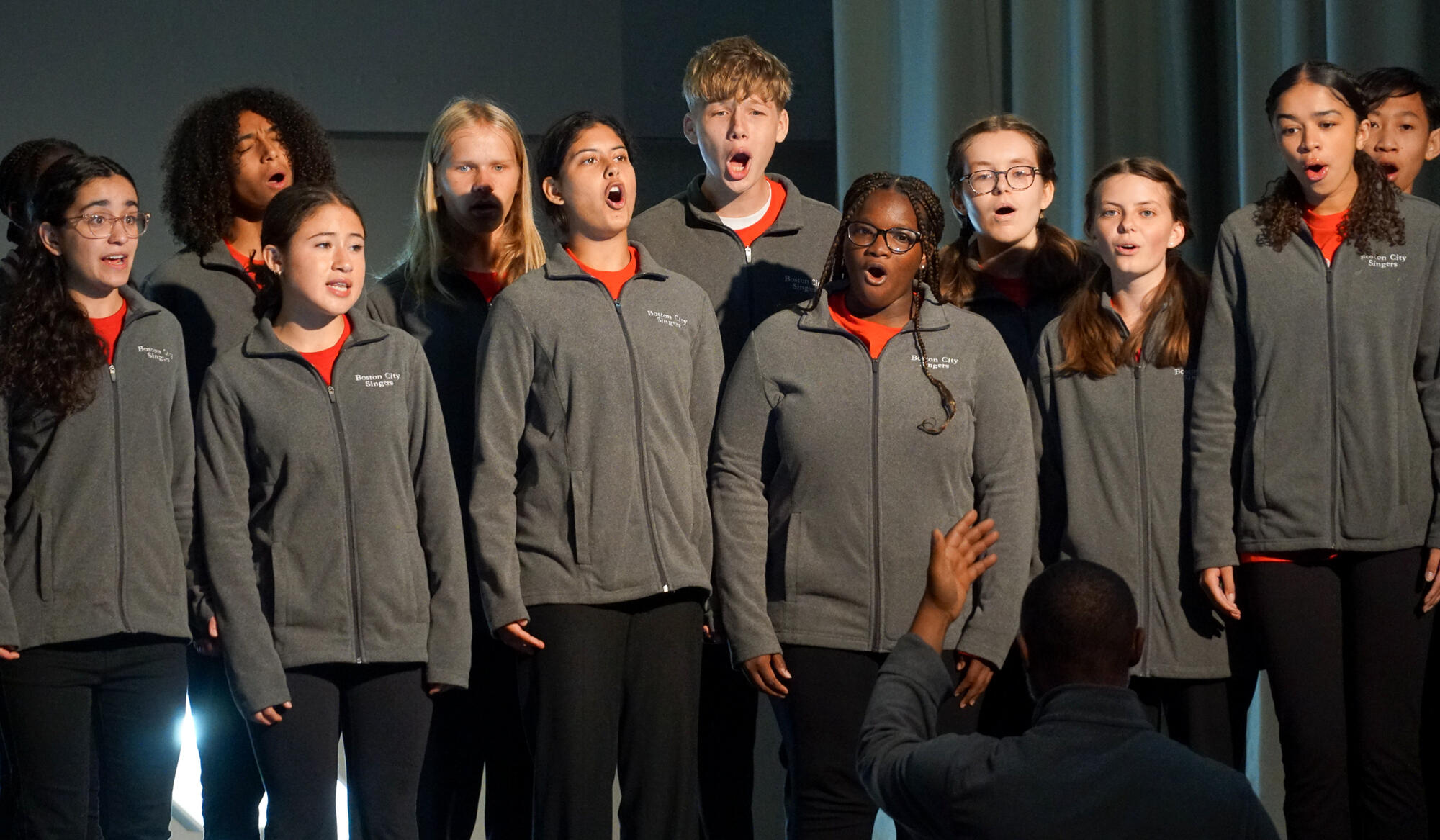 Group of middle school age children of varying ethnicities standing on stage wearing grey fleece jackets singing