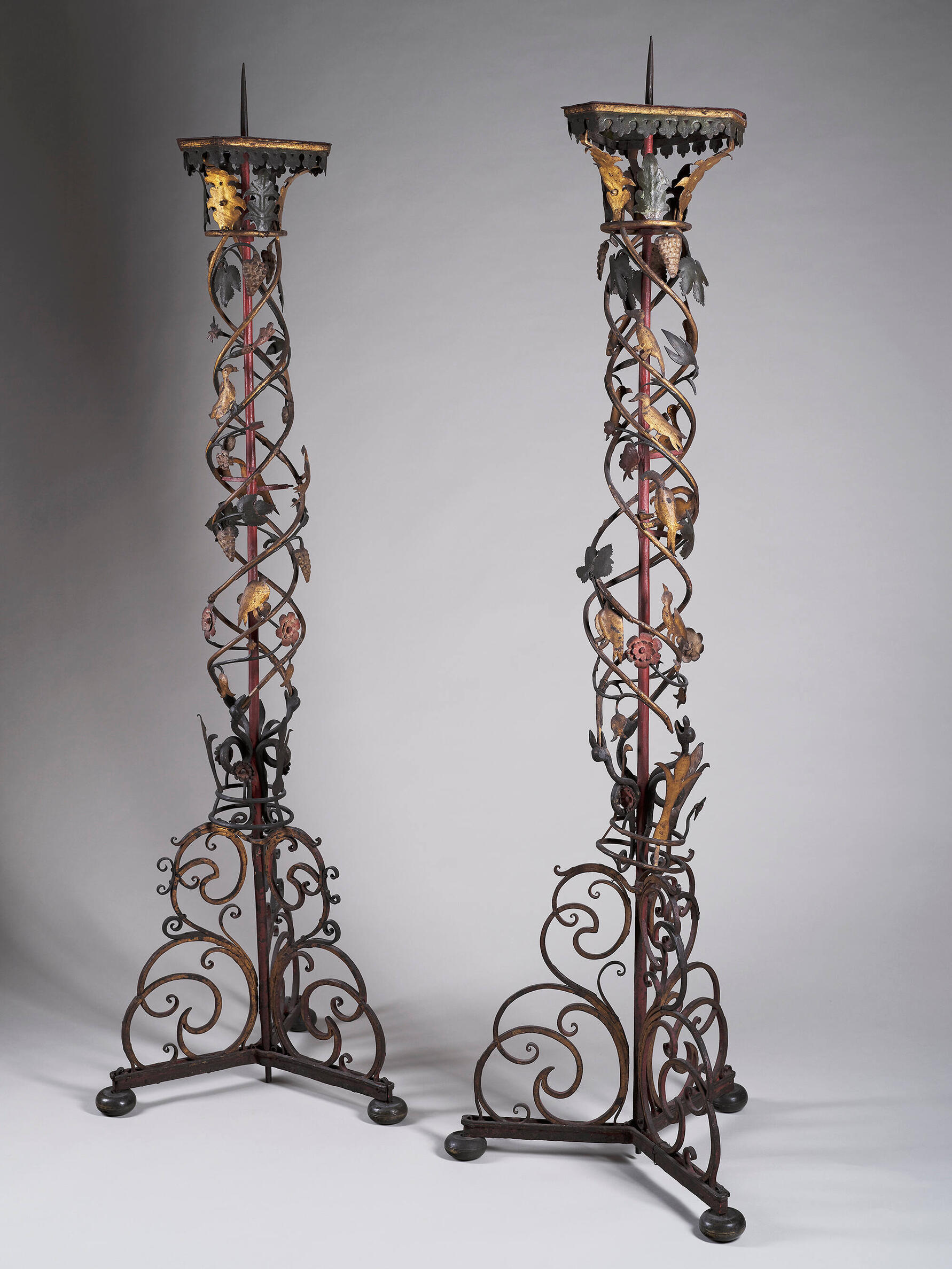 Two iron tall standing candlesticks with spikes, decorated with scrolling metalwork, gilded leaves, grapes, and birds. 