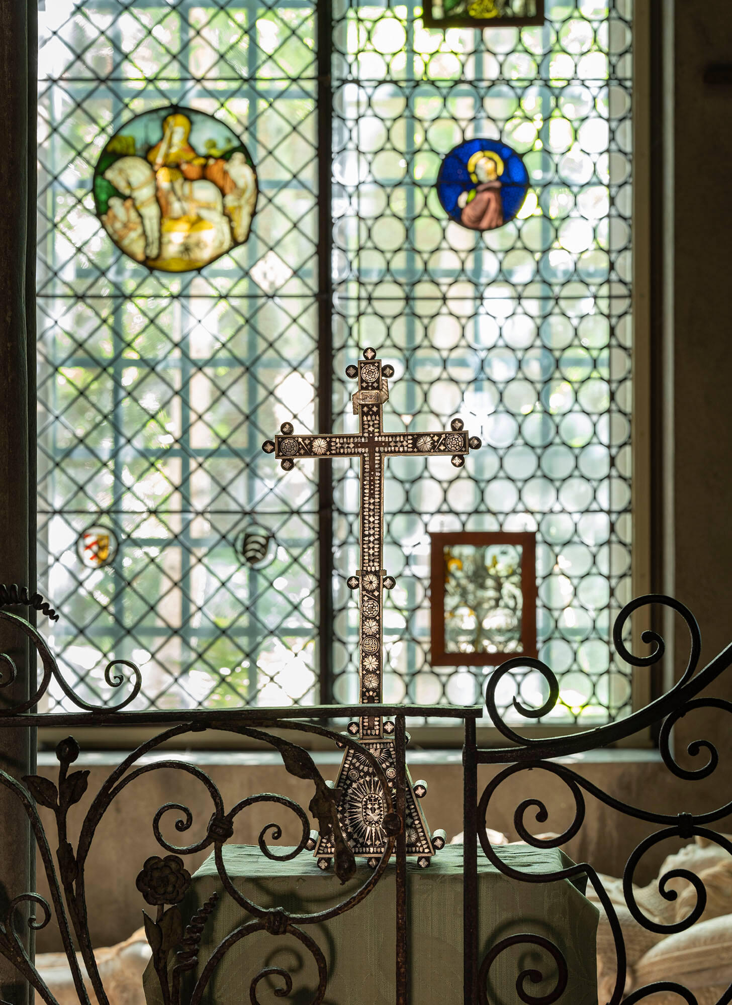 A wooden cross decorated with mother-of-pearl sits on a table in front of stained glass windows.