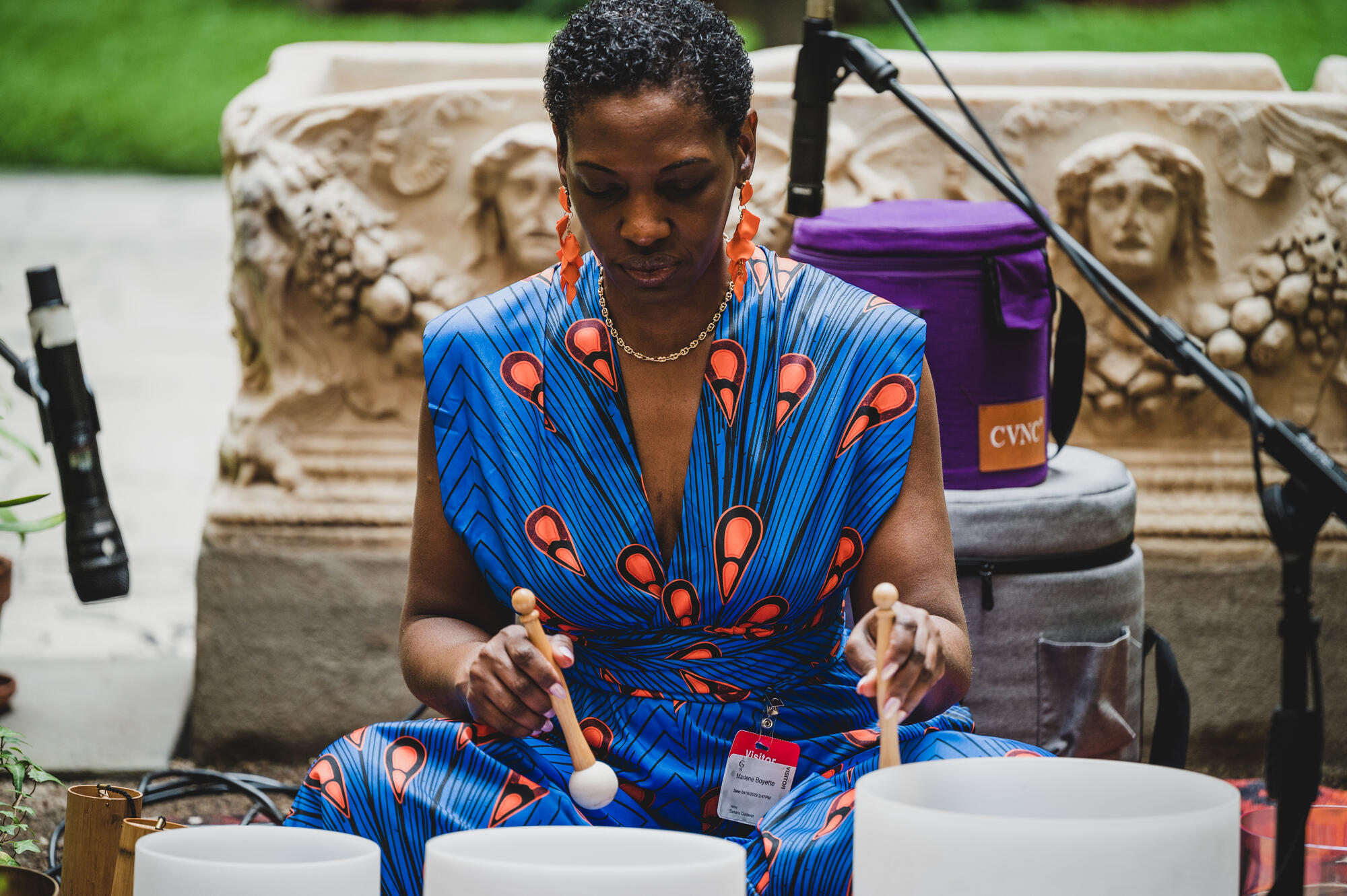 African American woman with short cropped hair wearing vivid blue and orange dress sitting on the ground leading a sound bath with glass containers. 