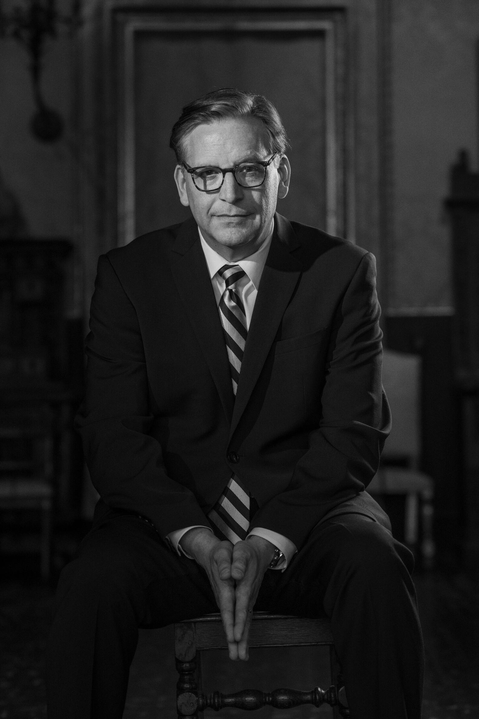 A black and white photo of a causcasian man sitting in a chair infront of an empty frame behind him wearing glasses, a suit and tie and looking directly at the camera. 