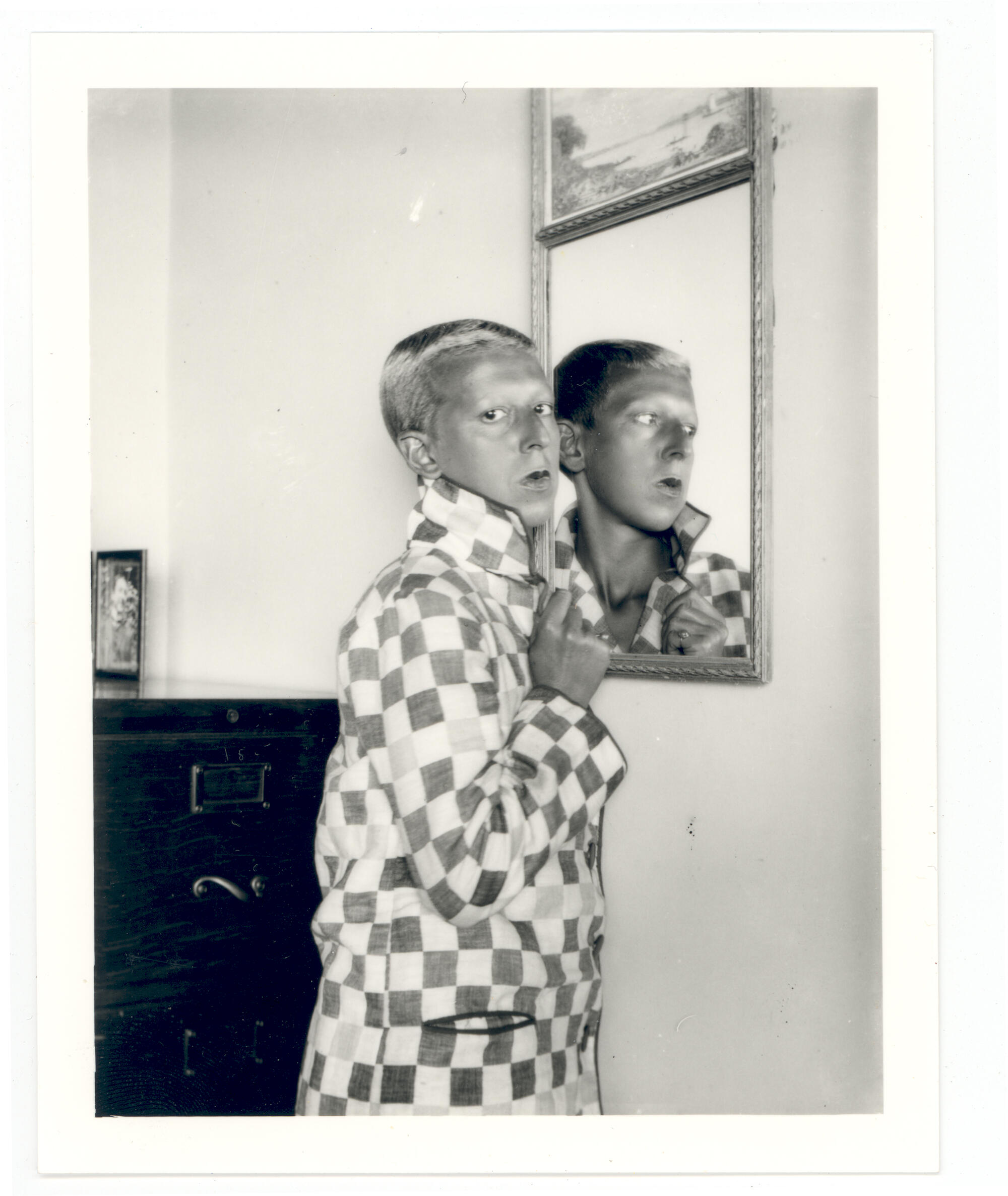 Black and white photo of a caucasian man with a blonde crew cut, wearing a checkerd jacket, standing in front of a mirror but turned to face the camera. 