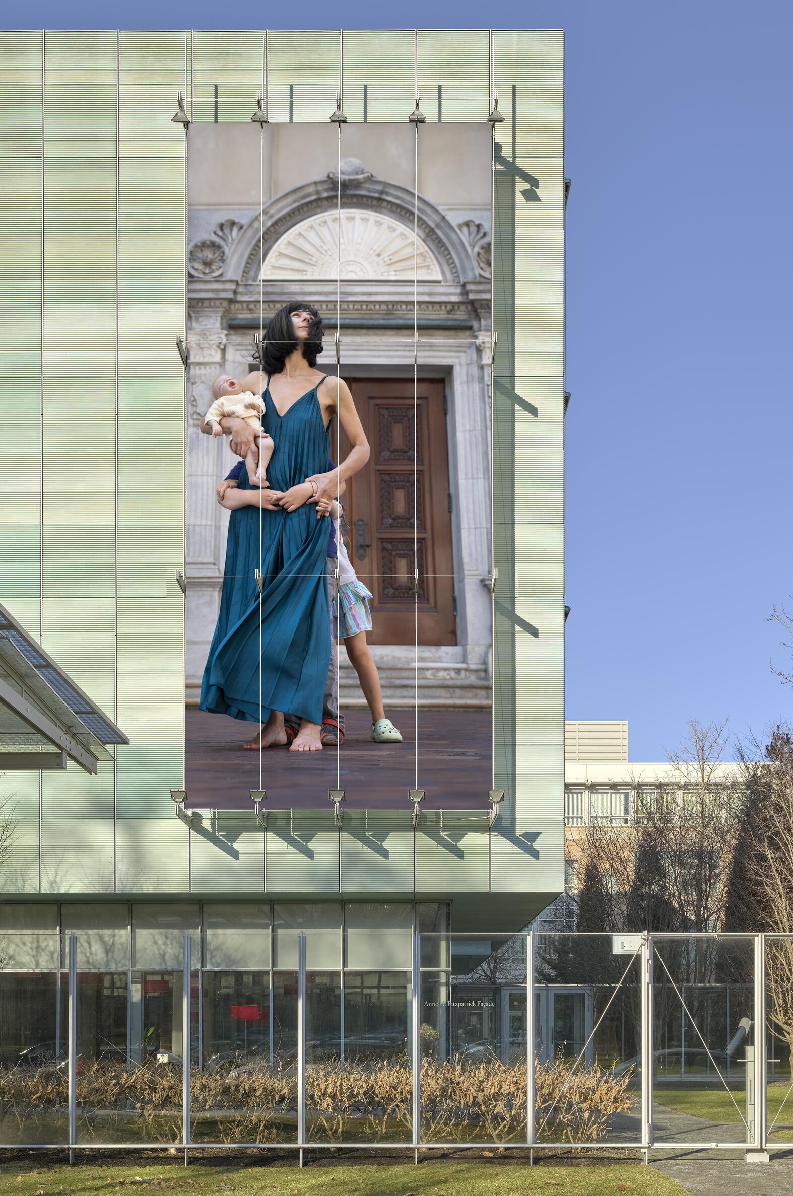 A woman in a sleeveless turquois long dress standing in a classical, stone building holding a baby doll with a small child hiding behind her. 