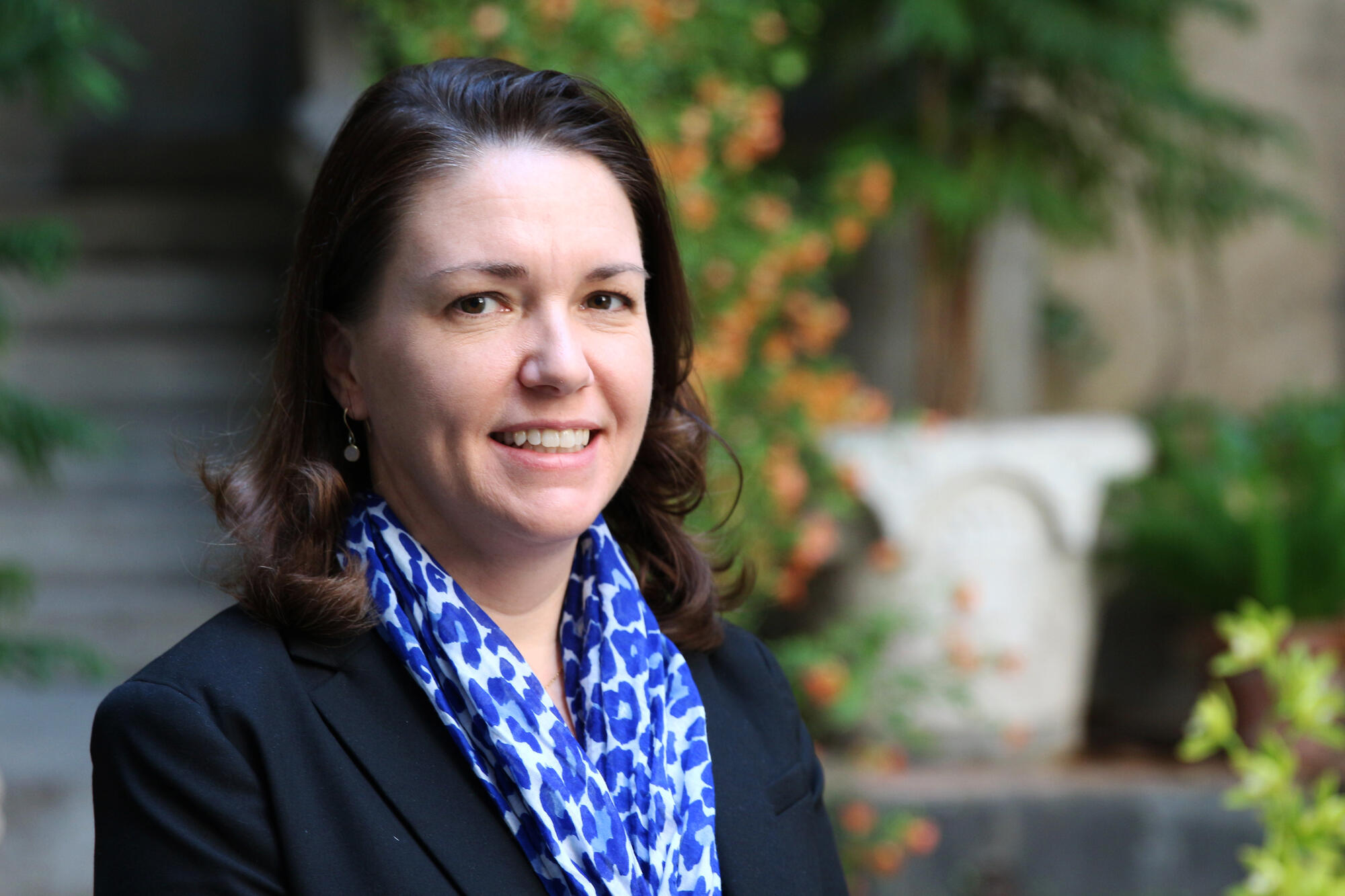 Caucasian woman with shoulder length dark brown hair wearing a suit jacket and silk scarf, in front vines of blooming nastruriums smiling at the camera. 