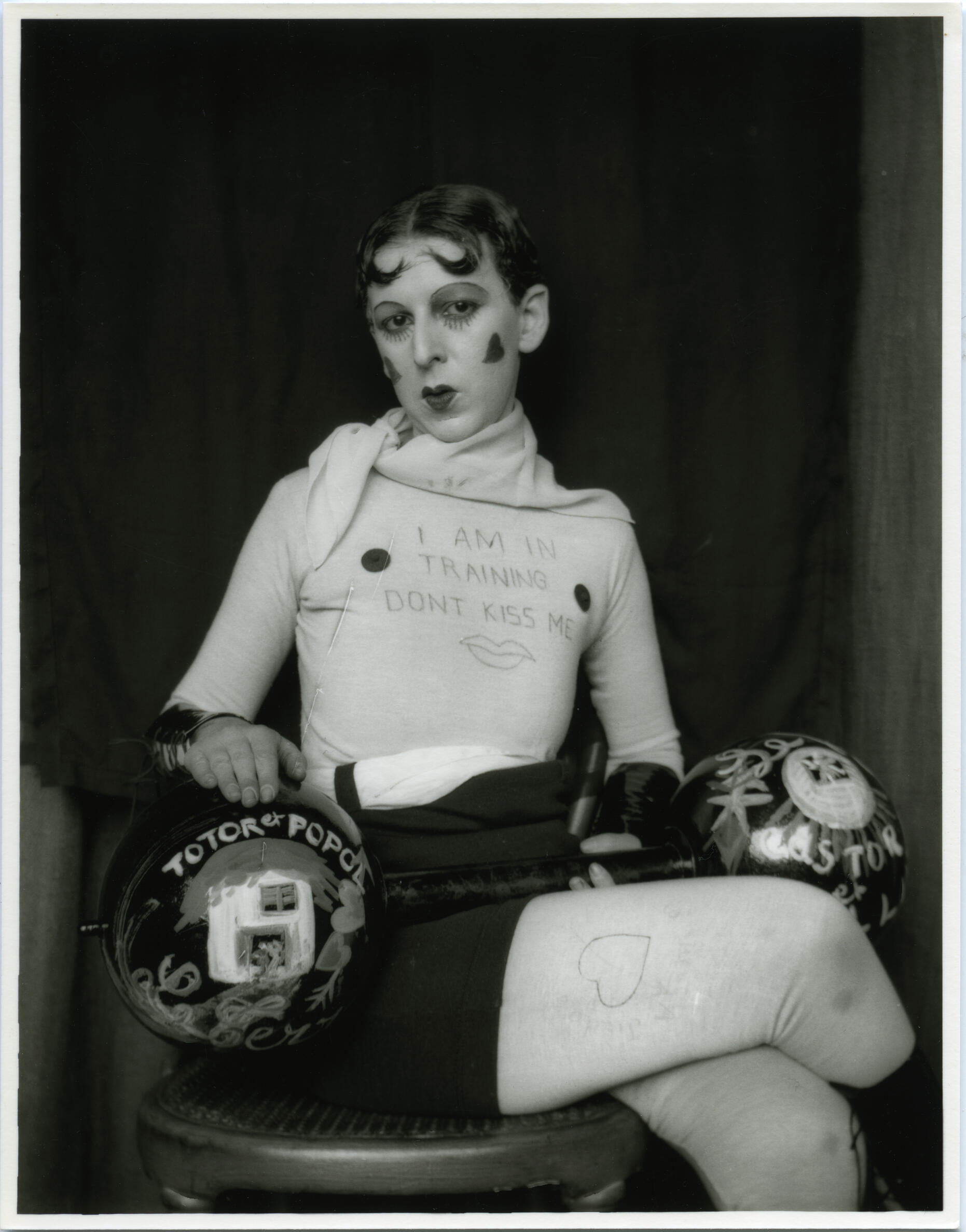 Black and white photograph of a man wearing a tight shirt with nipples drawn on and the words "I am in training don't kiss me" written on it.  He is wearing a short skirt with his hair slicked back with lipstick and rouge circles on his face. 