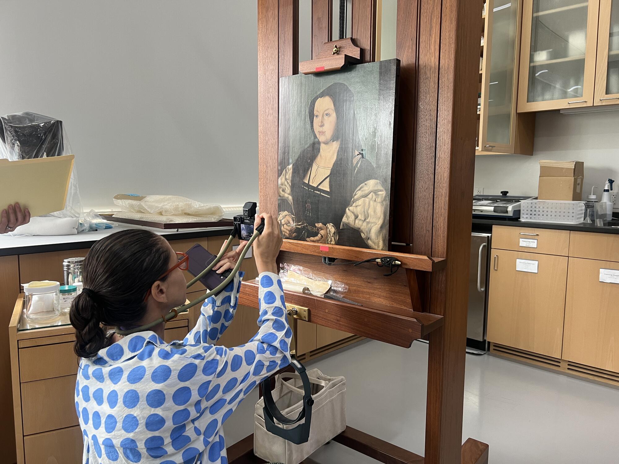 A woman kneeling on the floor taking a photo of a painting being cleaned in a laboratory like setting. 