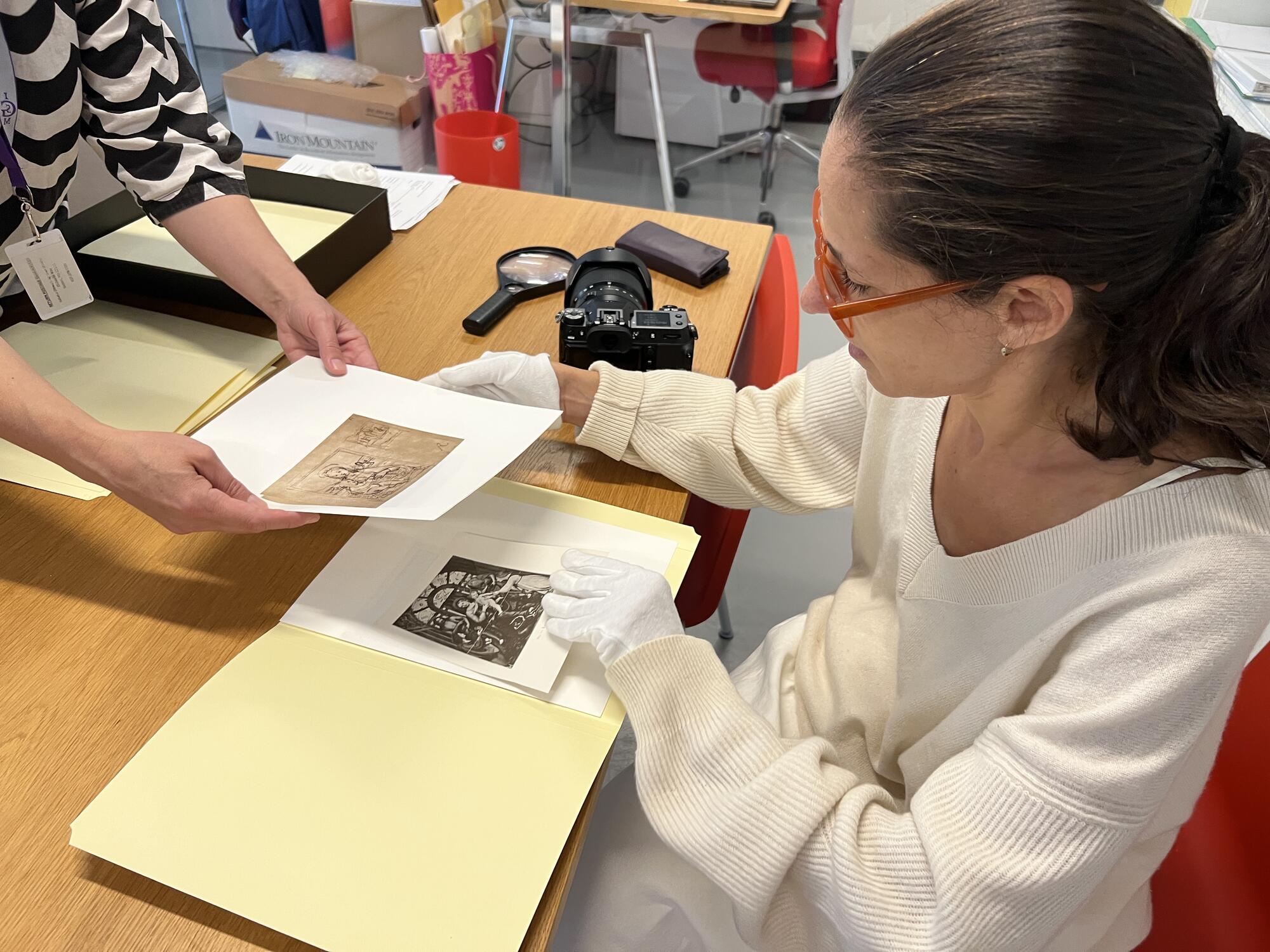 A woman sitting at a desk with white, cloth gloves on, examining old photographs. 