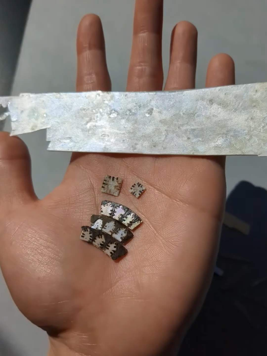 A strip of imitation mother-of-pearl and replacement pieces are laid out on a hand in the sun and rotated slightly to show the shift and reflection of the material. Underneath the strip, are mock-ups of different shapes, matching the mother-of-pearl on the object.