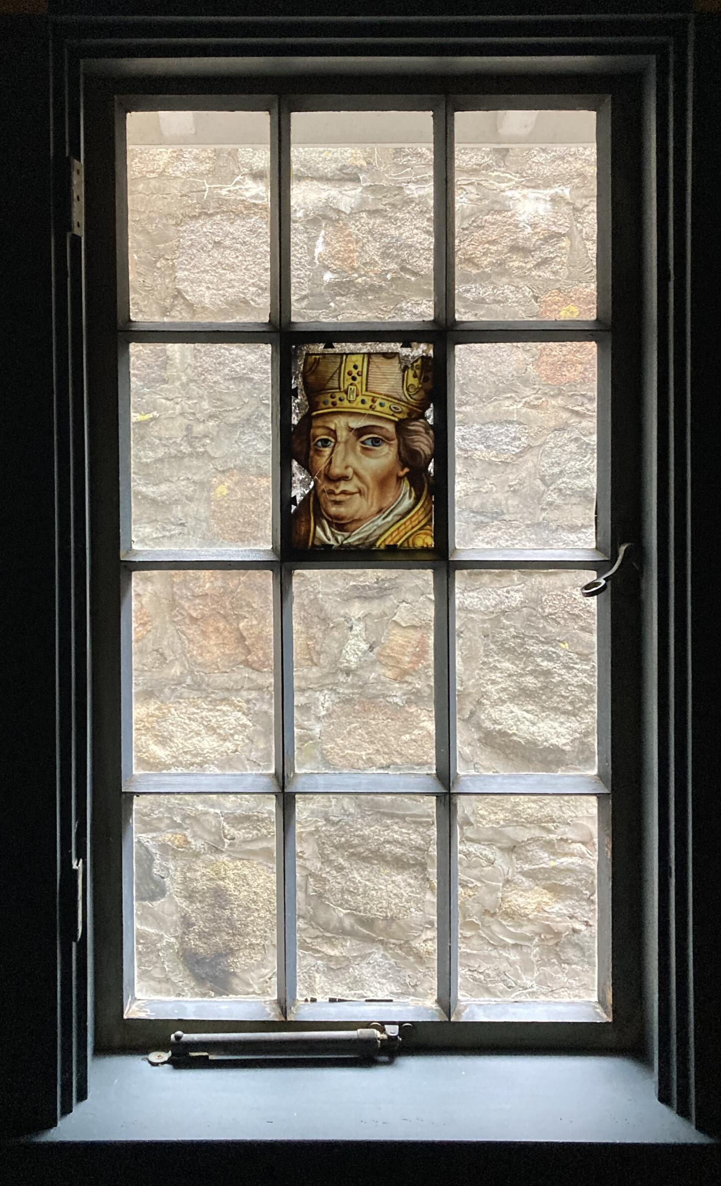 A twelve-paned window with a stained glass pane of a bishop’s head with a lazy eye.