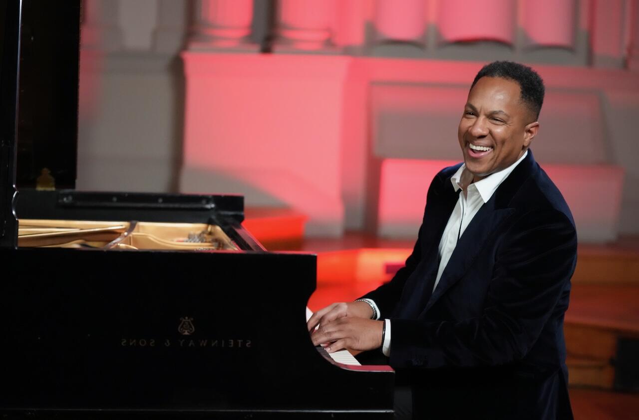 African American man wearing a dark suit and white shirt playing the piano with a big smile on his face. 