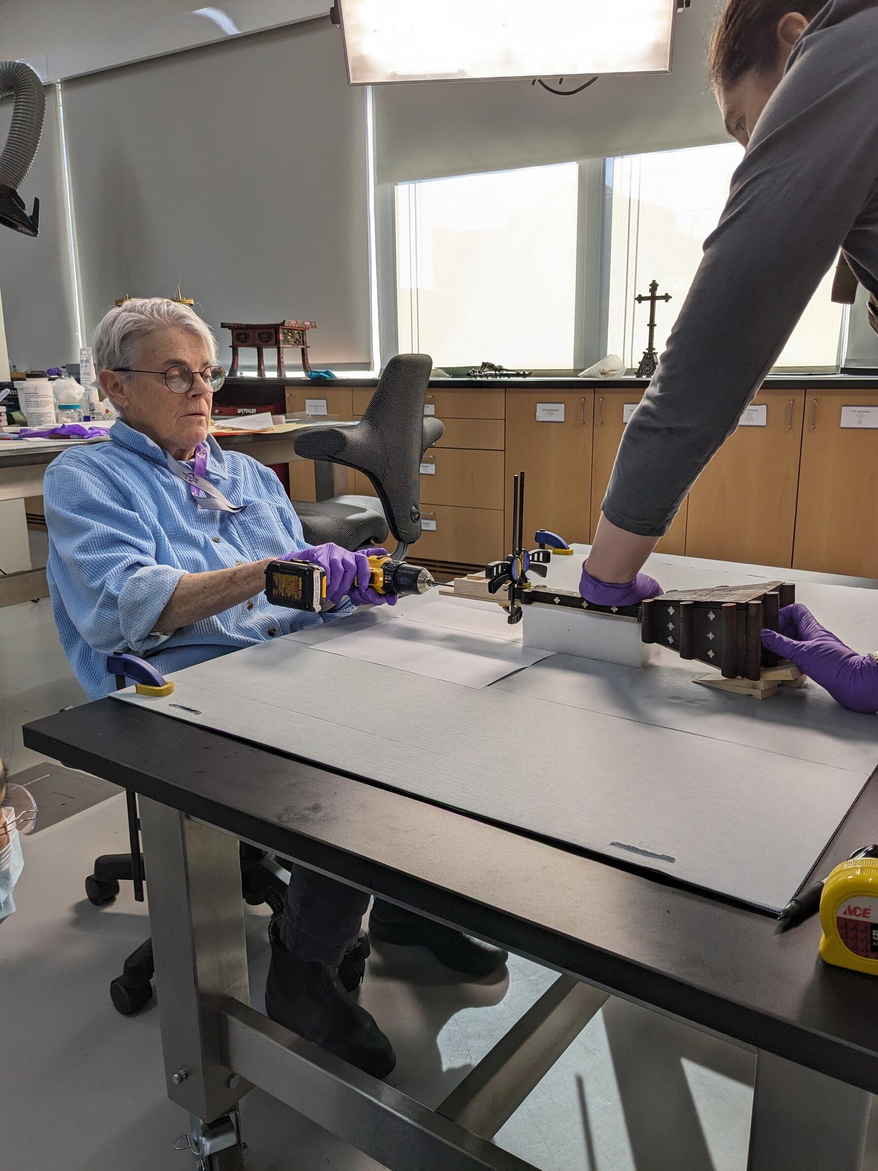 The cross is laid down horizontally with support as contract conservator (Melissa Carr) and Objects Conservator, Jessica Chloros, drill into the bottom half of the cross as part of the mechanical repair. 
