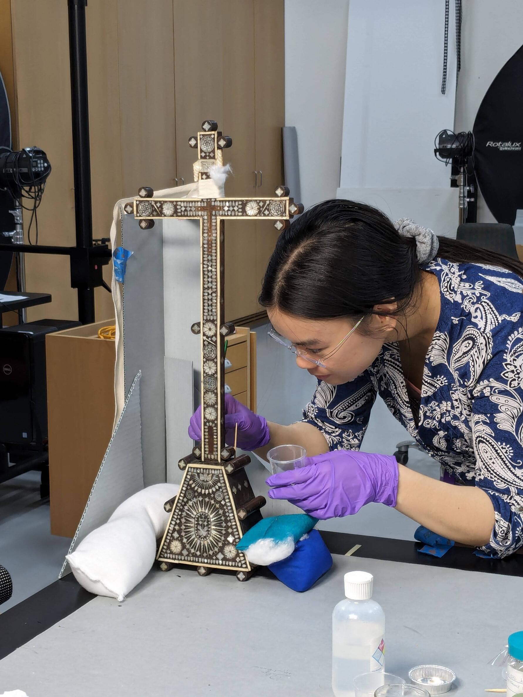  A conservator is applying adhesive to the base of the cross which is tied to a temporary support so it does not move while the adhesive sets. 