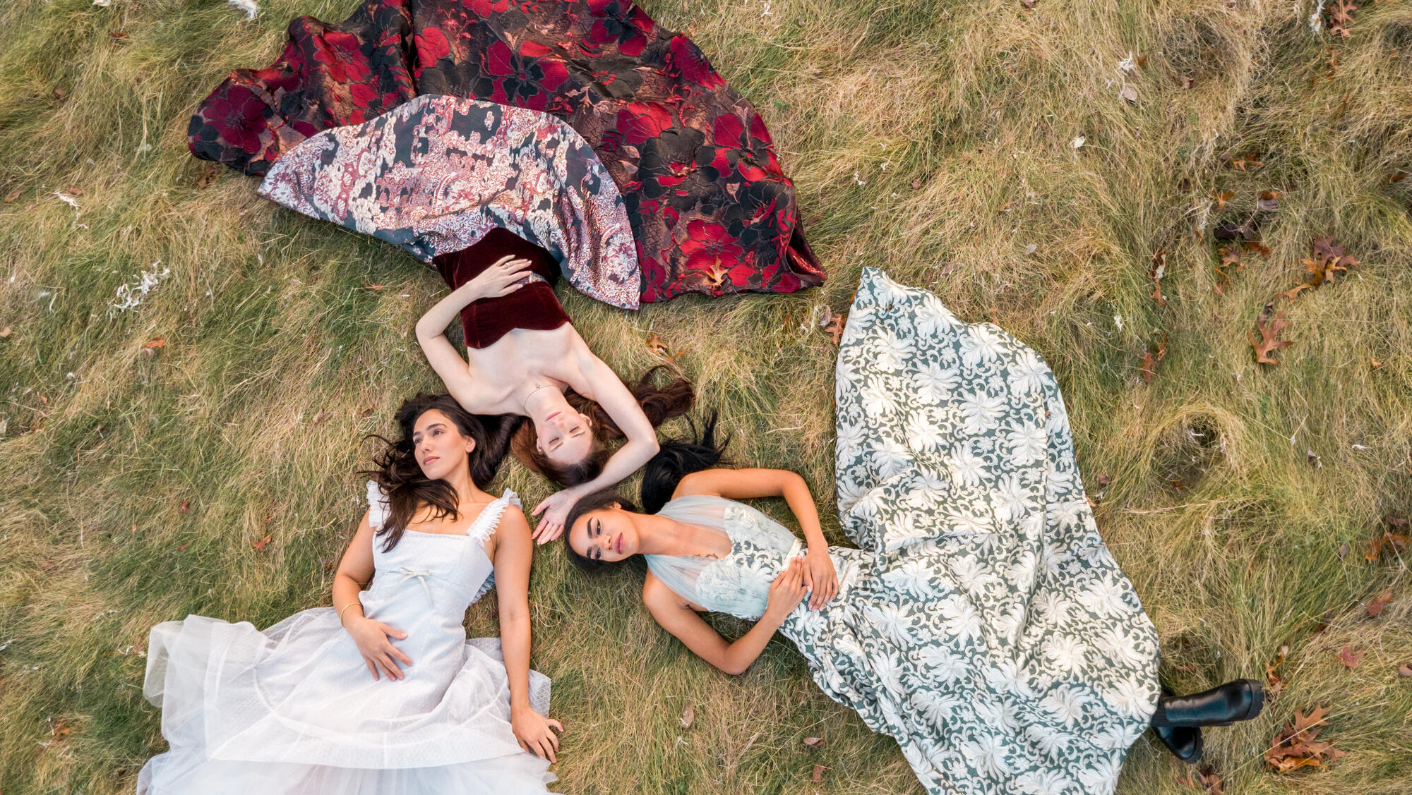 Three young, caucasion woman wearing long flowing dressing lying on the ground with their heads almost toucing in the middle, looking up to the sky. 