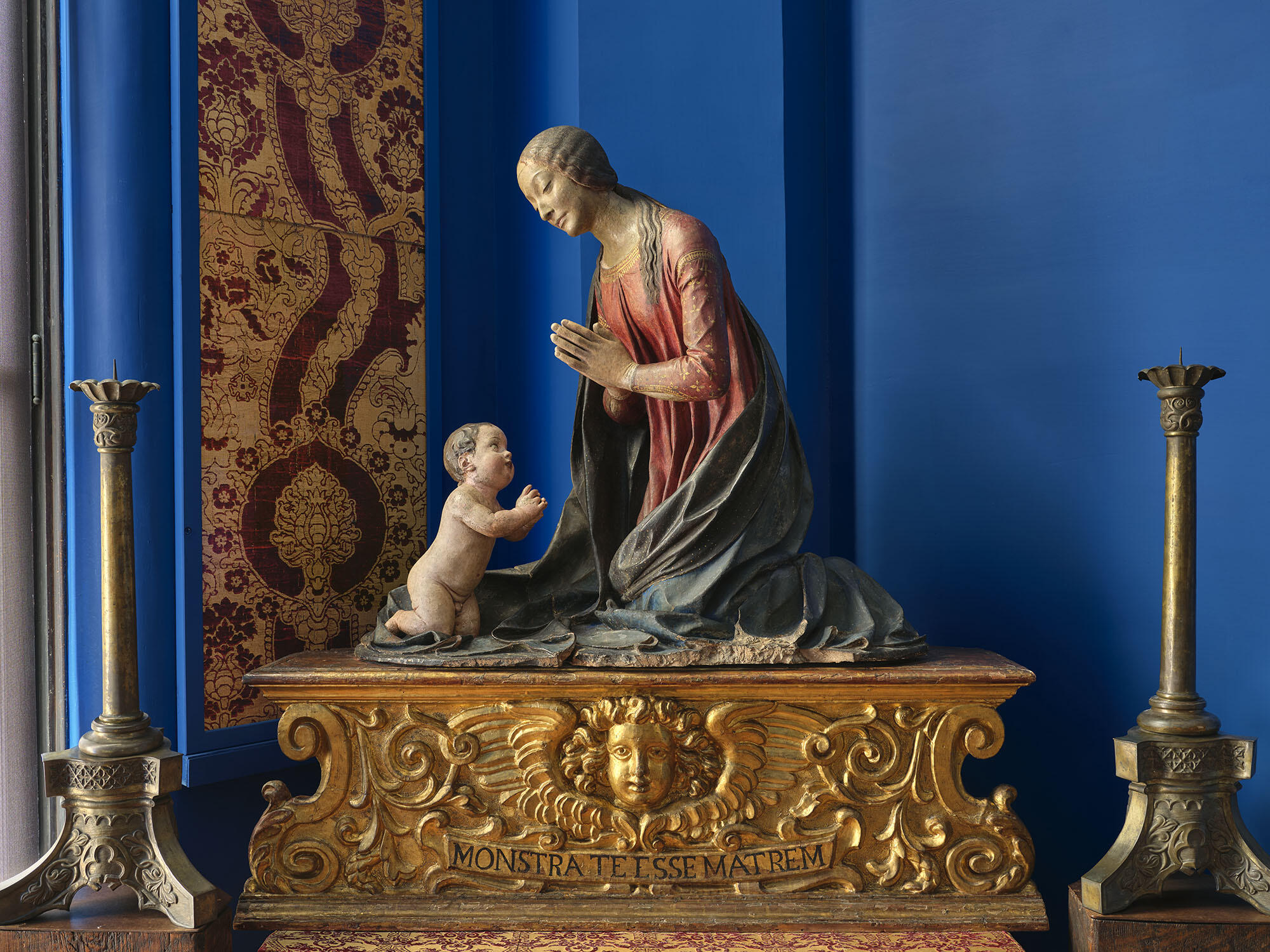 An almost life-sized painted and gilded terracotta sculpture of kneeling figures of the Madonna and Child holding their hands in prayer while looking at each other in mutual adoration.