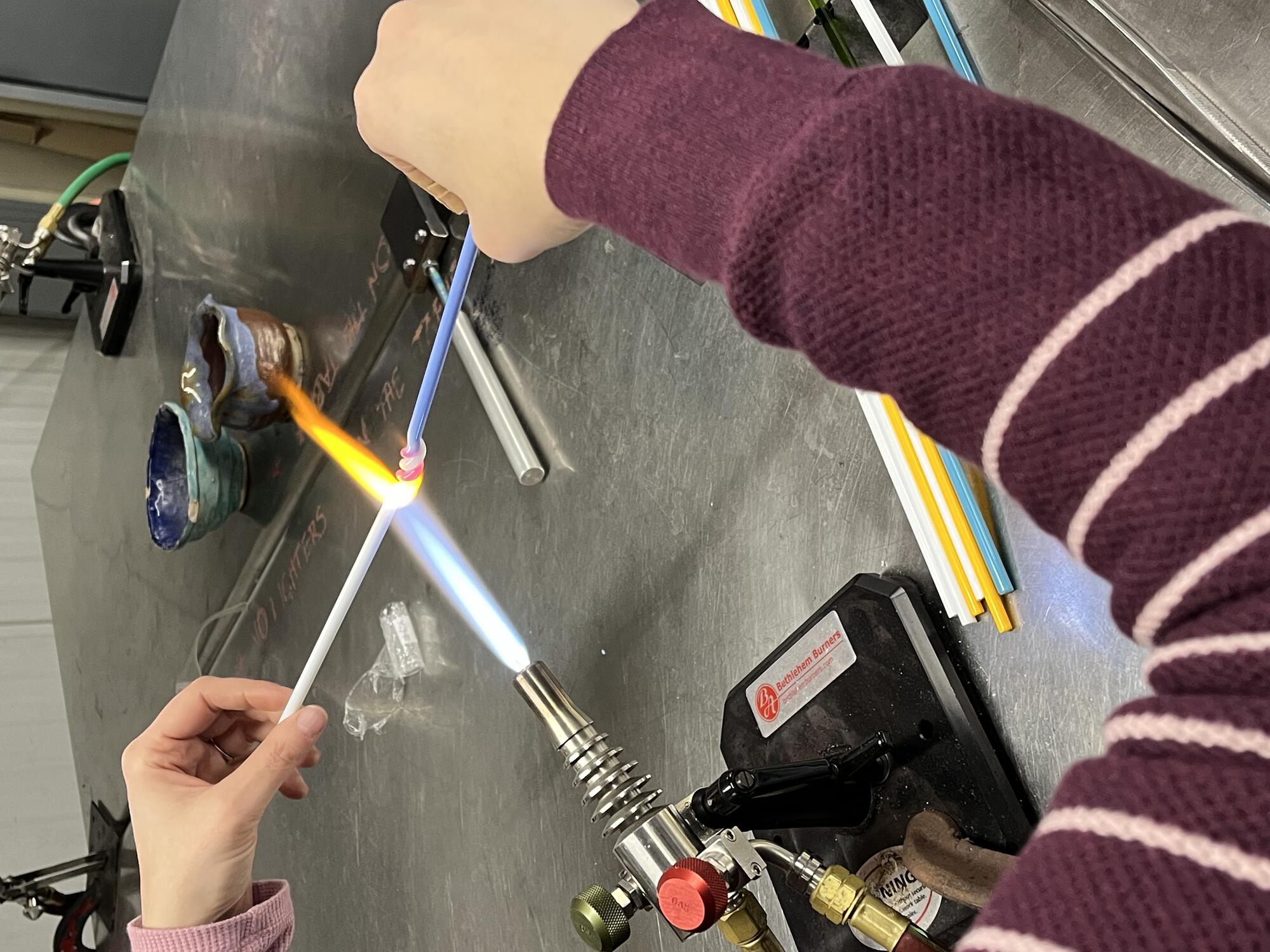 Detail of a person holding a rod of blue glass in their right hand and a rod of white glass in their left hand over a torch that is mounted to the table in front of them.  The rods are getting twisted together in the middle as they soften over the flame.  