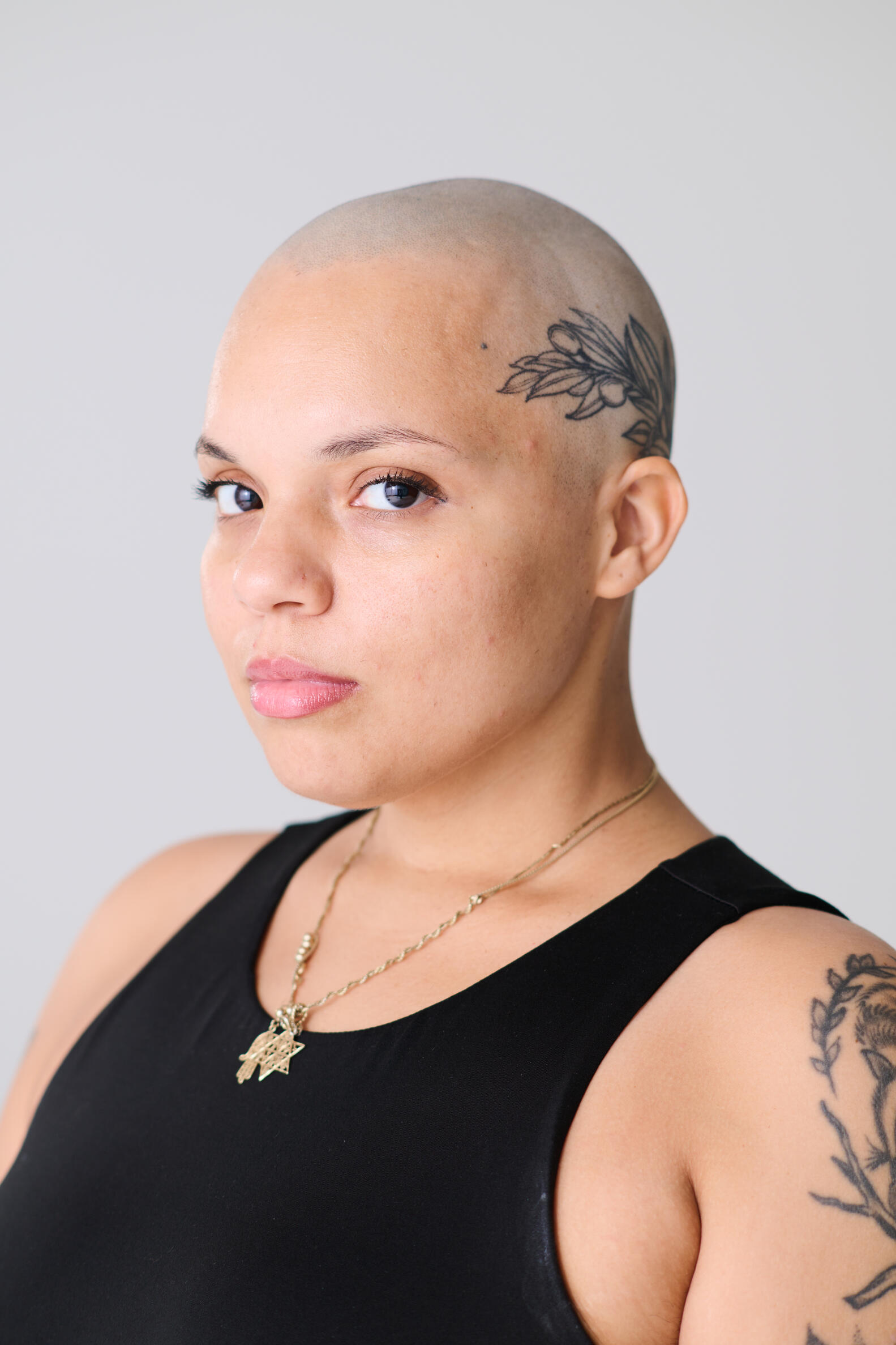 headshot of person with flower tattoo on the side of their head wearing a black tank top and gold necklace