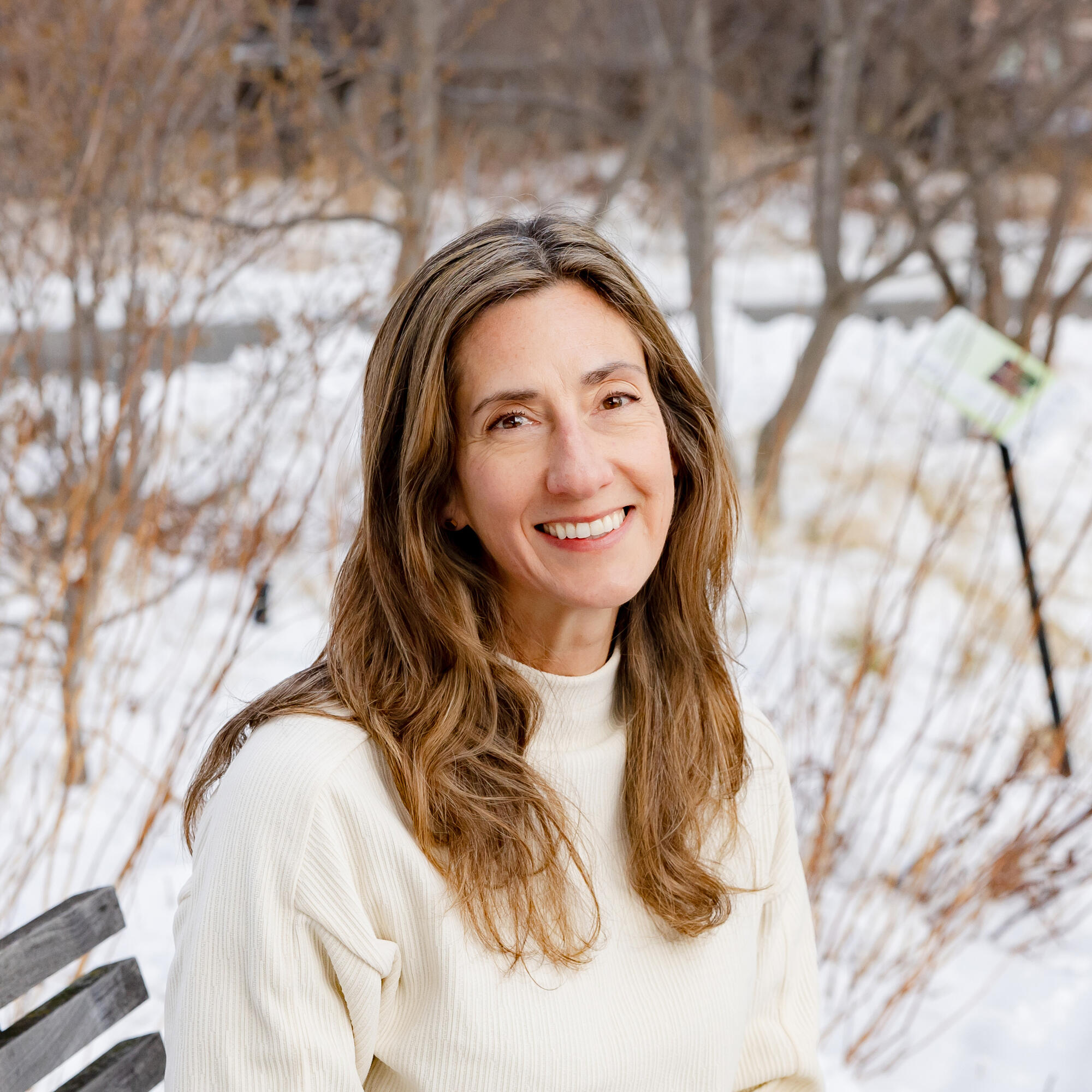 Headshot of woman with brown hair sitting on a park bench with snow behind her