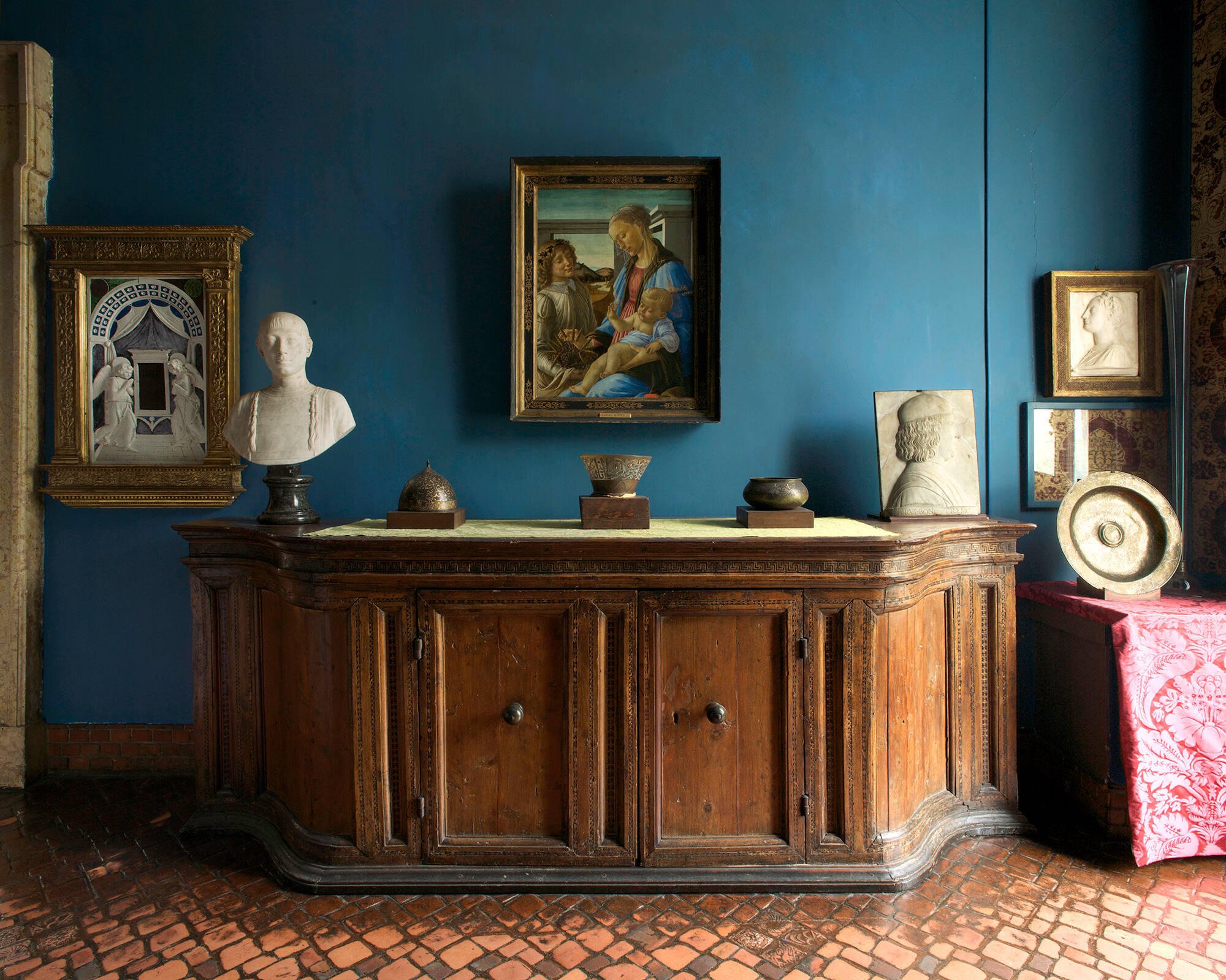 A flared neck and rim of a glass oil lamp on top of a wooden credenza flanked by an Islamic metal bowl and helmet. The oil lamp fragment is opaque and has a white support at its base.