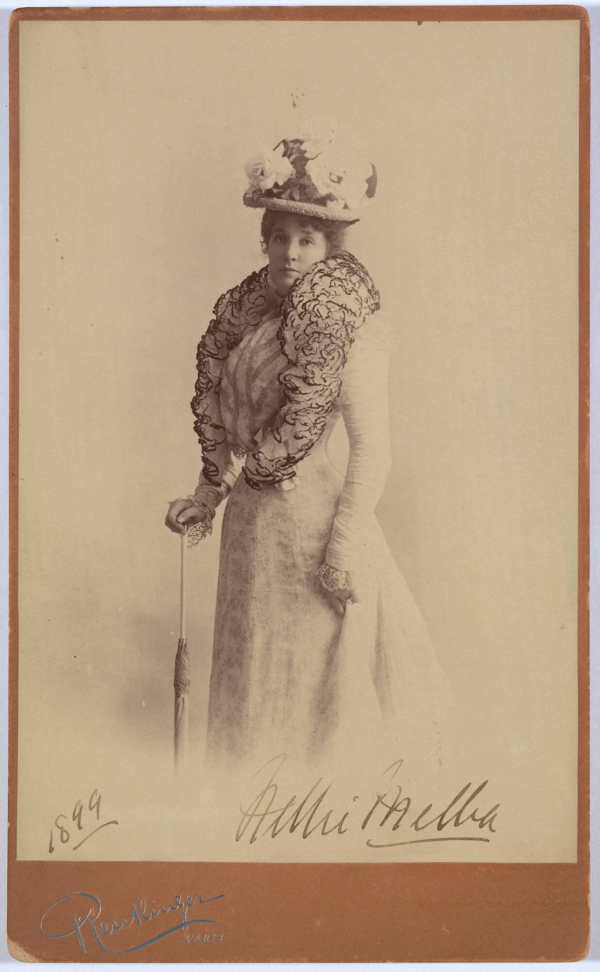 A black and white photograph of a white woman wearing a light colored dress with long sleeves and lace cuffs and a hat with large flowers on it.