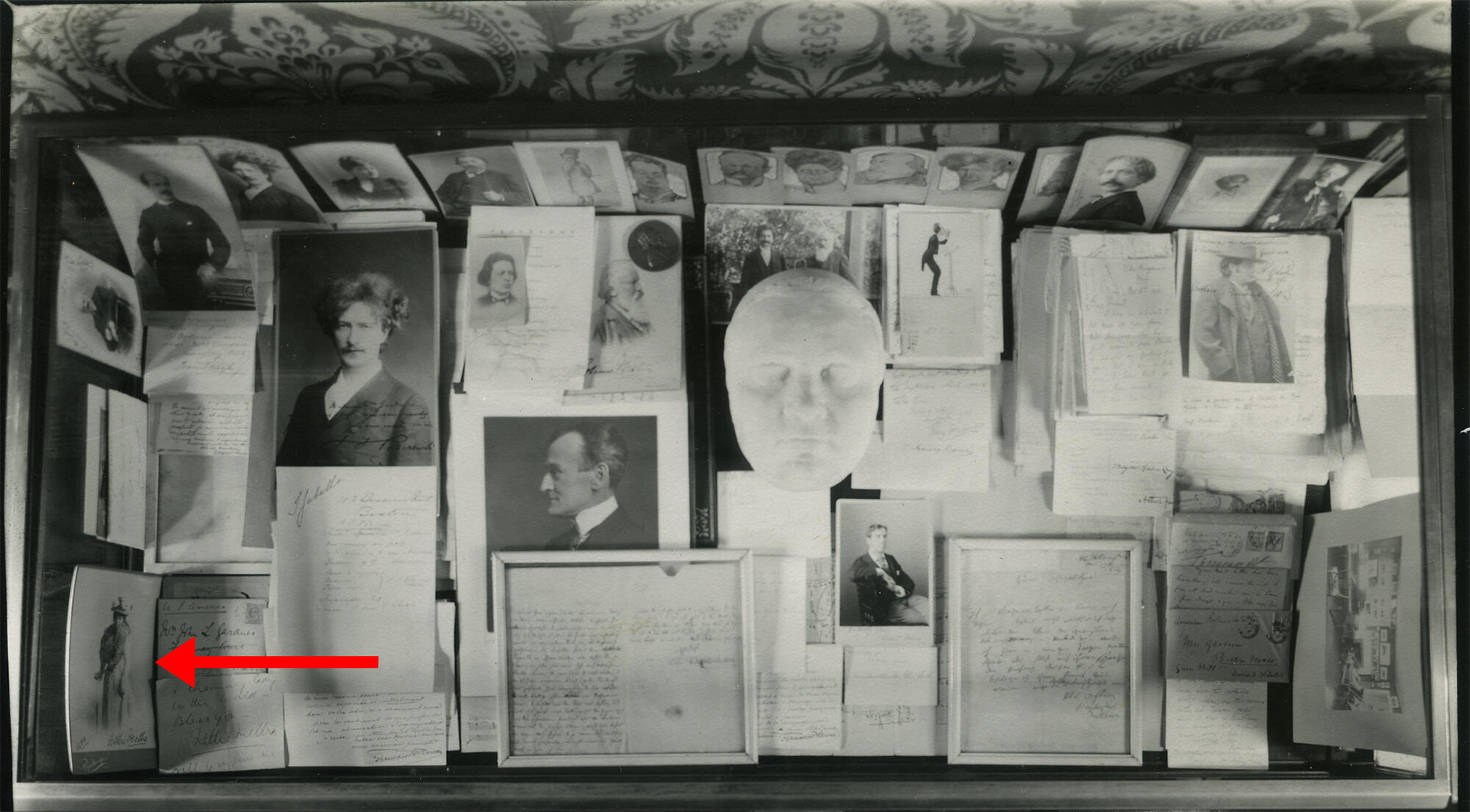 A black and white photograph of a glass topped display case filled with letters and photographs related to musicians. A red arrow indicates the photograph of Nellie Melba.