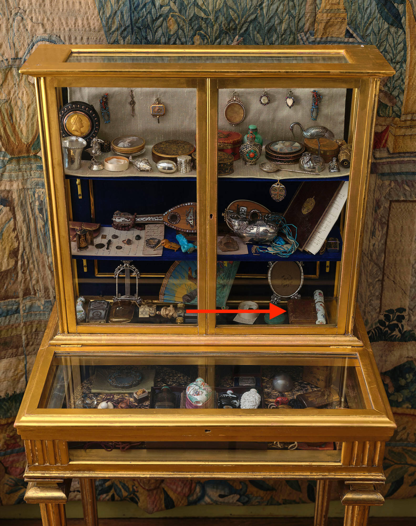 A glass display case containing many objects. A red arrow points to a brown leather case.