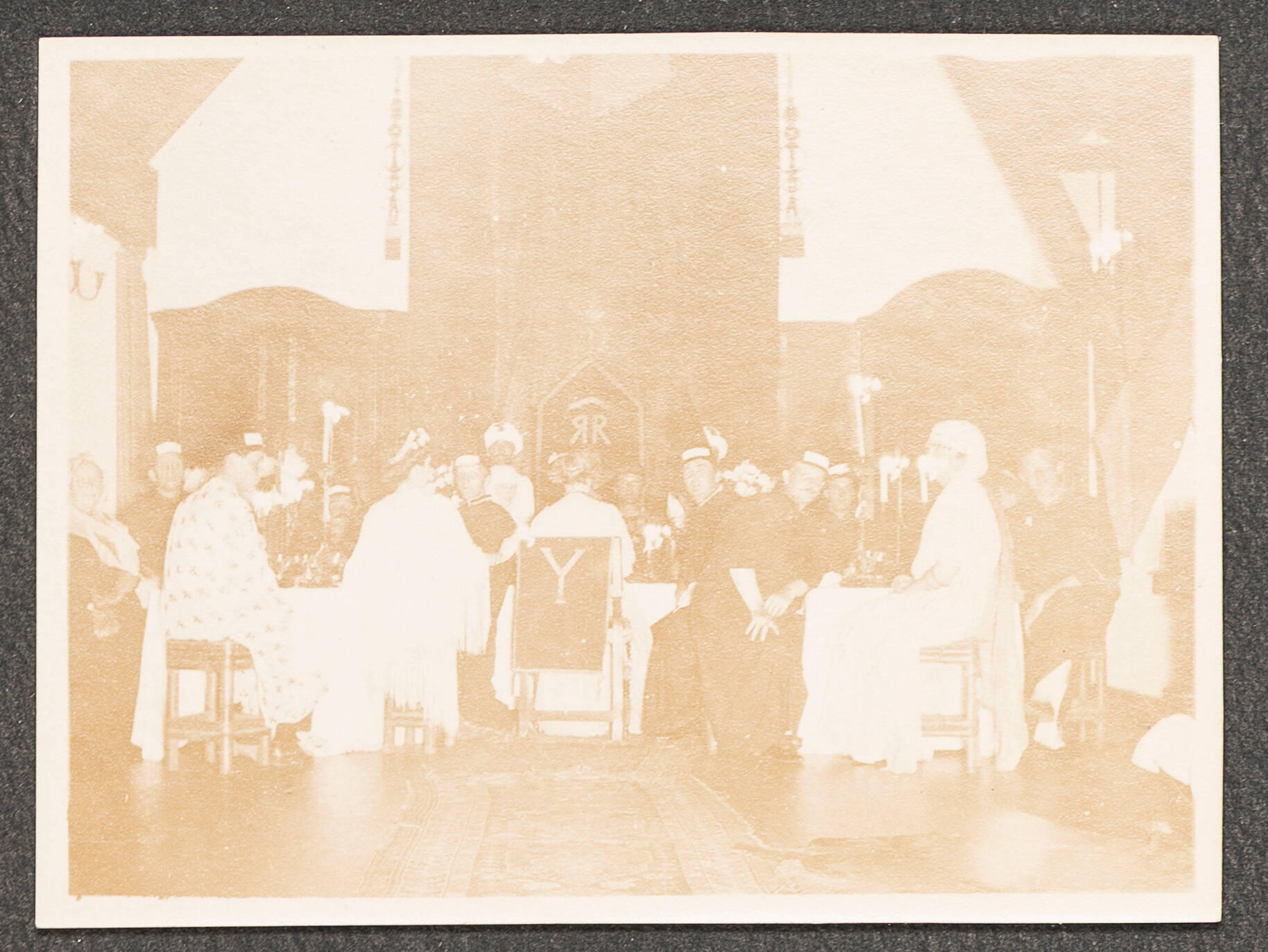 A black and white photograph of a dinner party at a table where the guests are dressed in costume. Isabella Stewart Gardner sits on a throne with the letter Y on it.